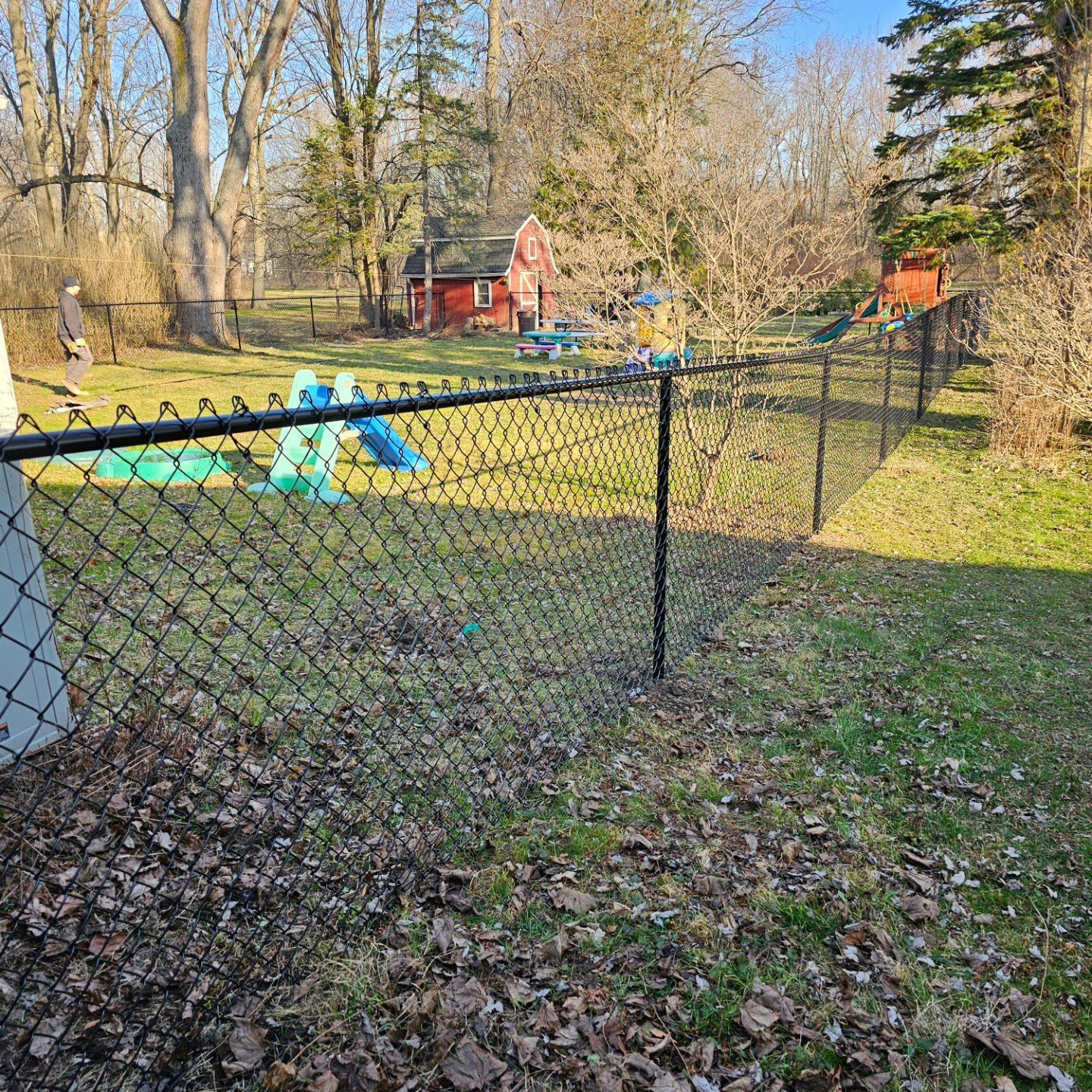 Chain-link fence around a leaf-strewn yard with a small blue playset and red shed in the background