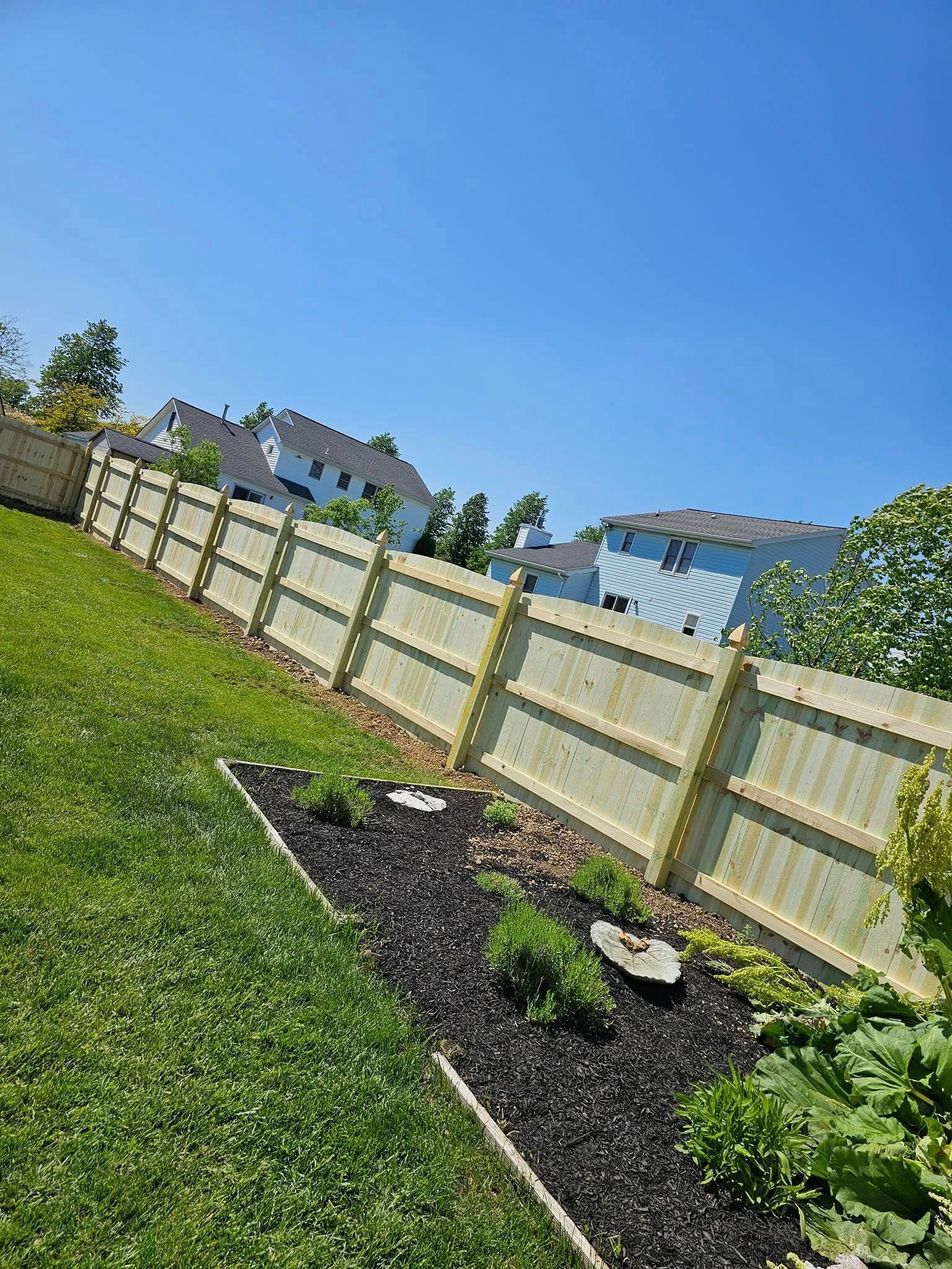 Sunny backyard with a slanted fence, green lawn, and a small mulched garden bed with young plants.