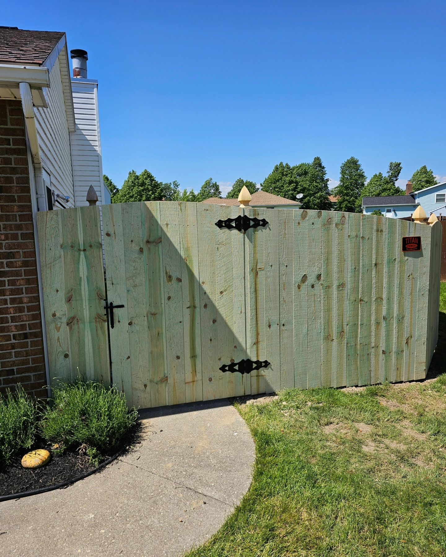 Weathered green wooden gate beside a house, opening onto a curving concrete path and grassy yard under a blue sky