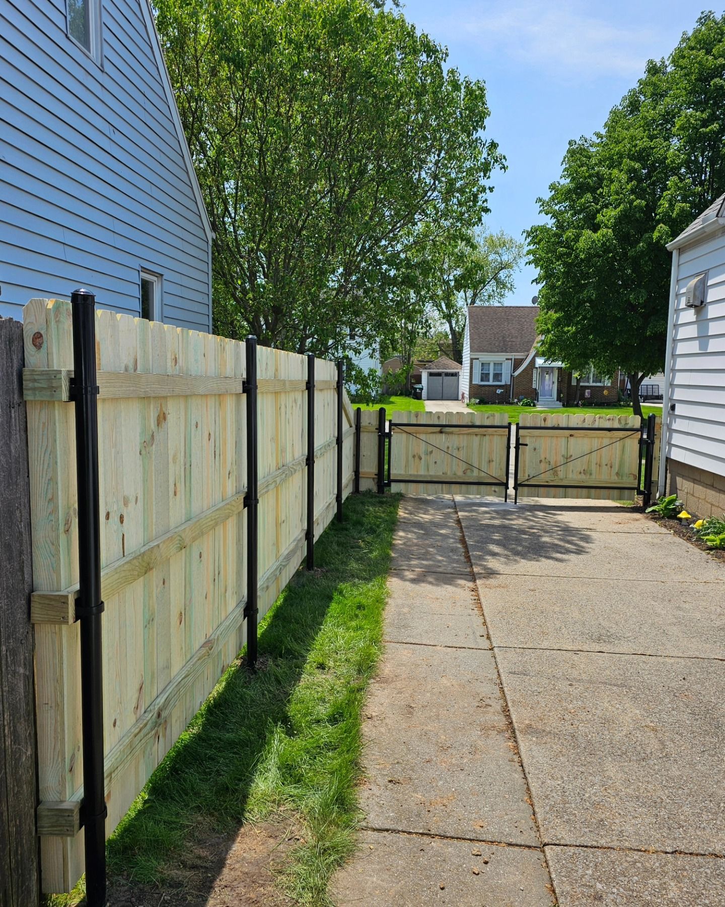 Driveway between blue and white houses with wooden fences and a gate under a sunny sky