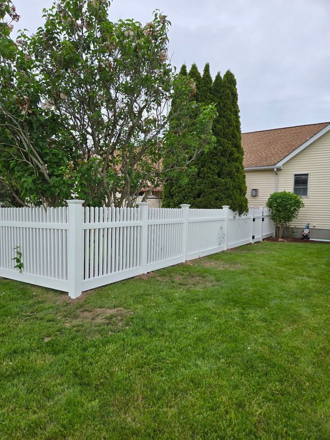 White picket fence along a grassy yard beside trees and a house under overcast skies