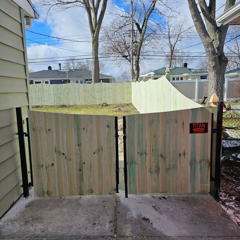 Wooden backyard gate with black hinges between concrete driveway and fenced yard under bare trees