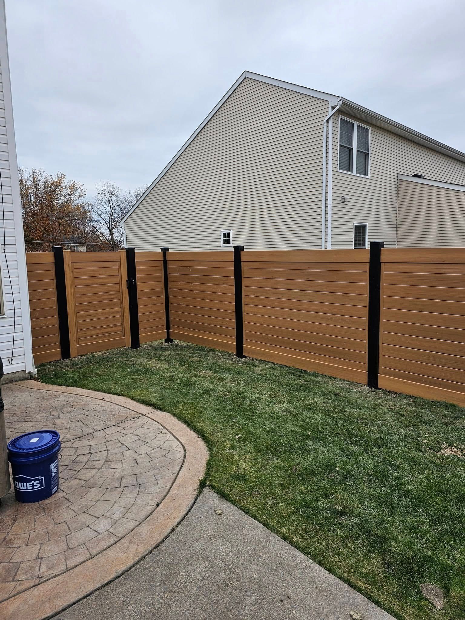Backyard with curved wooden fence, grass, patio stones, and a beige house under cloudy skies