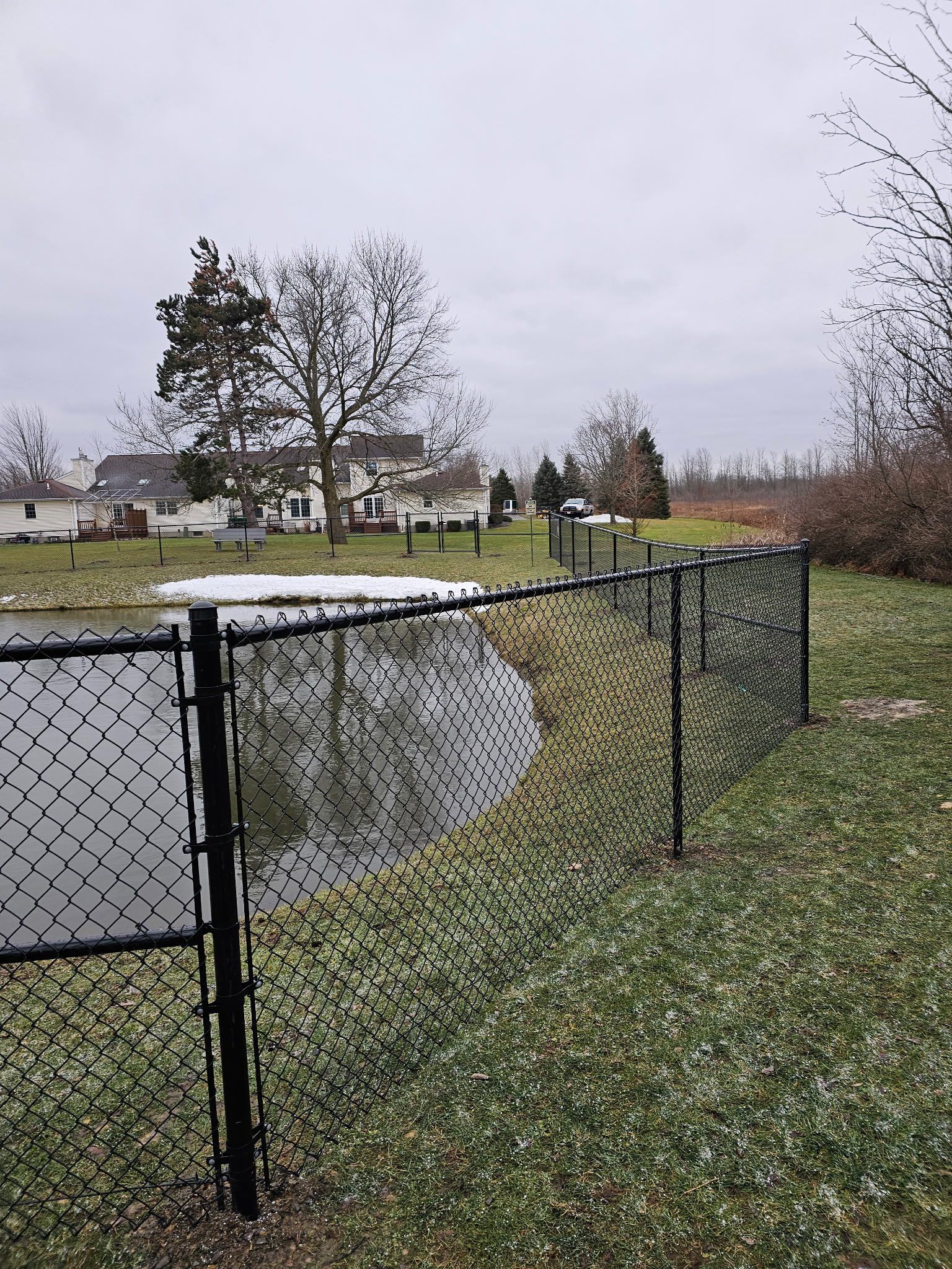 Chain-link fence beside a snowy pond in a grassy park under an overcast sky