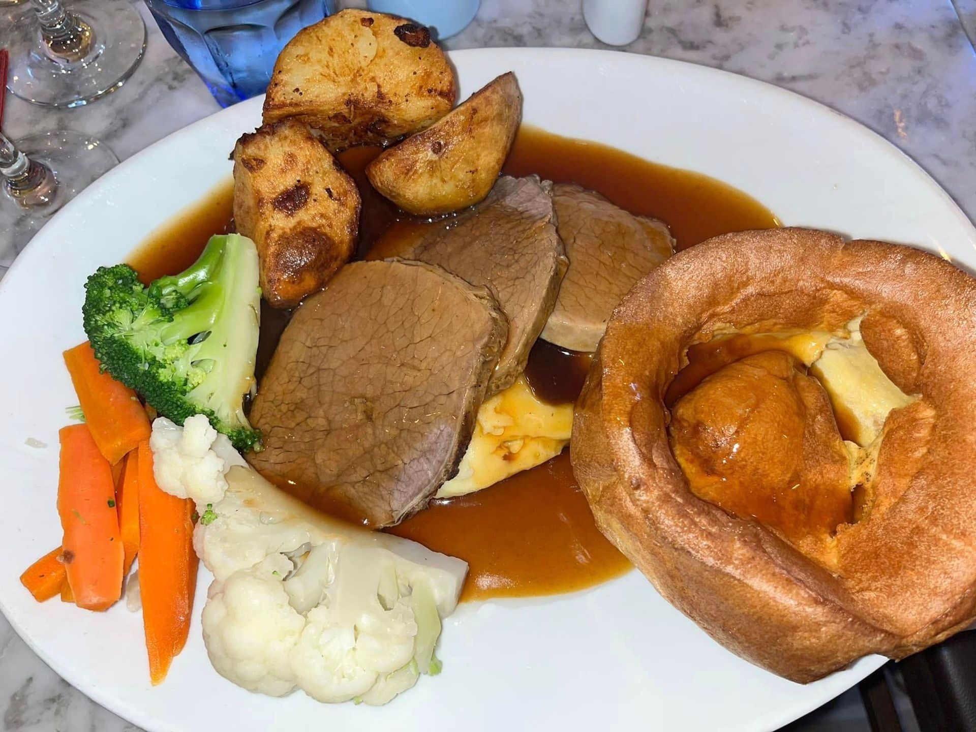 A classic Sunday roast dinner with beef slices, gravy, roast potatoes, a Yorkshire pudding, broccoli, and carrots.