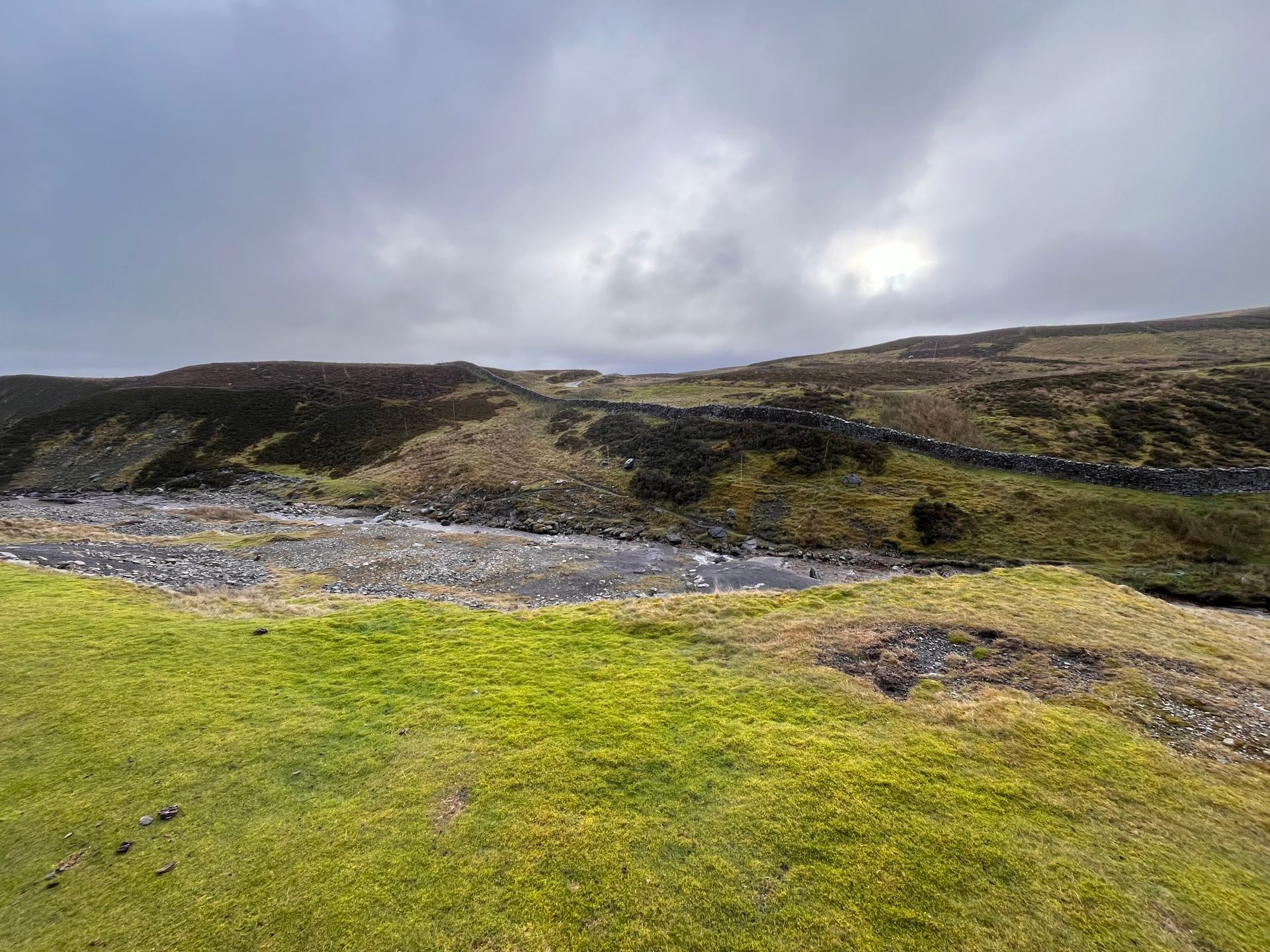 A river running through a grassy hillside with a cloudy sky in the background.