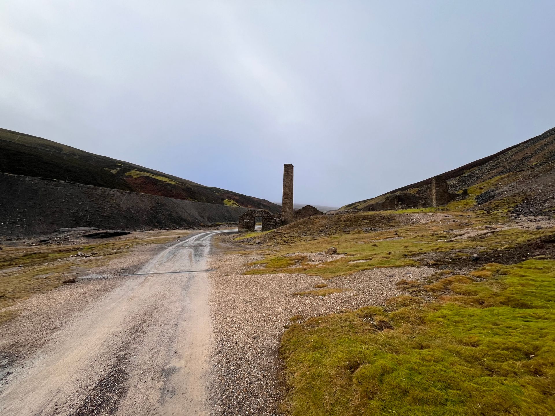 A dirt road going through a grassy field with a chimney in the background.