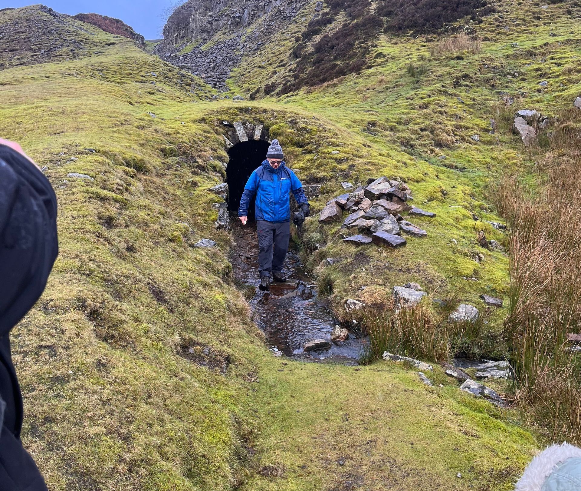 A person in a blue jacket walks out of a small, stone-arched tunnel in a mossy, hilly, outdoor landscape.