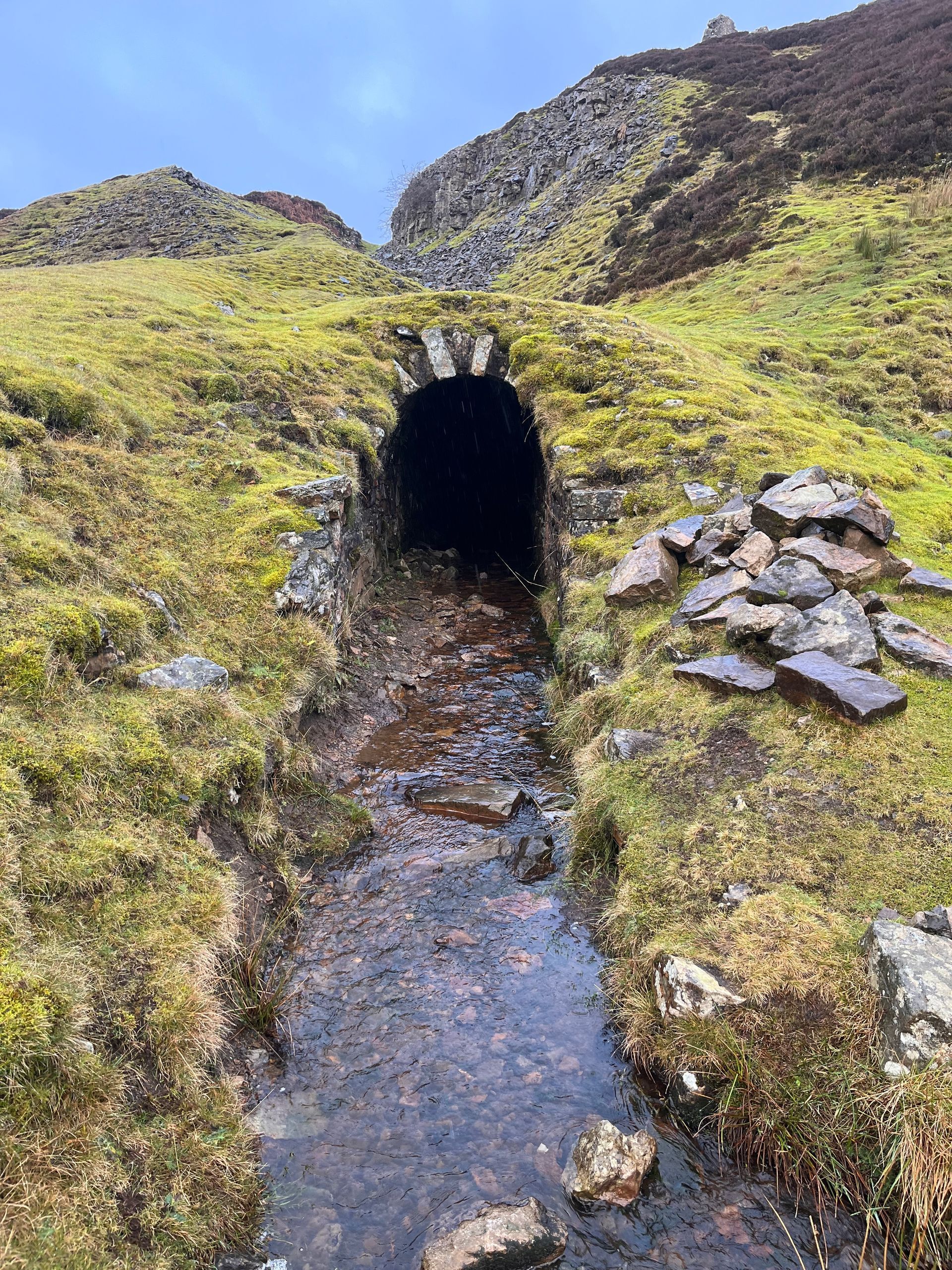 A small tunnel in the middle of a grassy field with a stream coming out of it.
