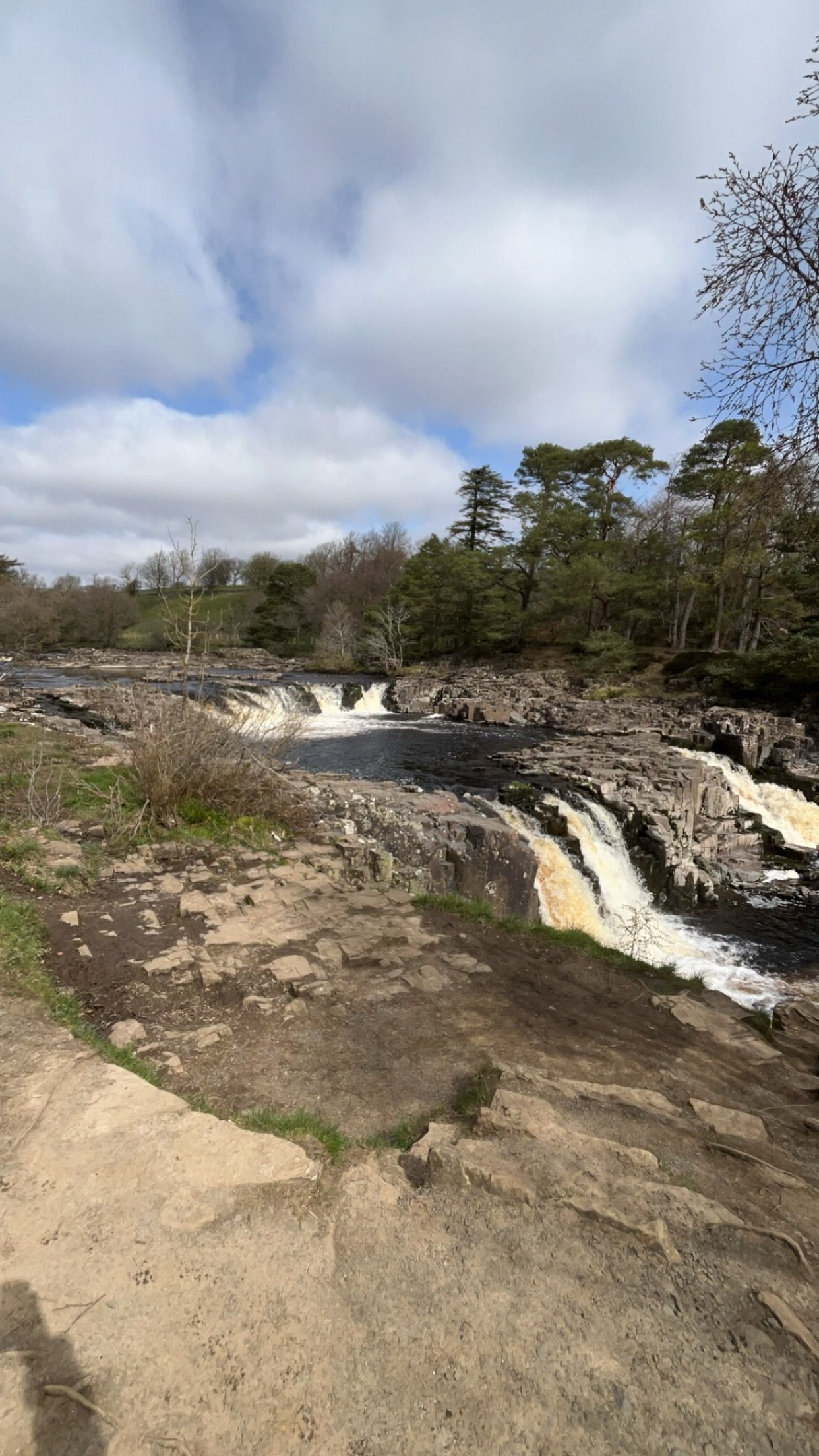 A tiered waterfall flows over rocky cliffs into a pool, surrounded by trees under a partially cloudy blue sky.