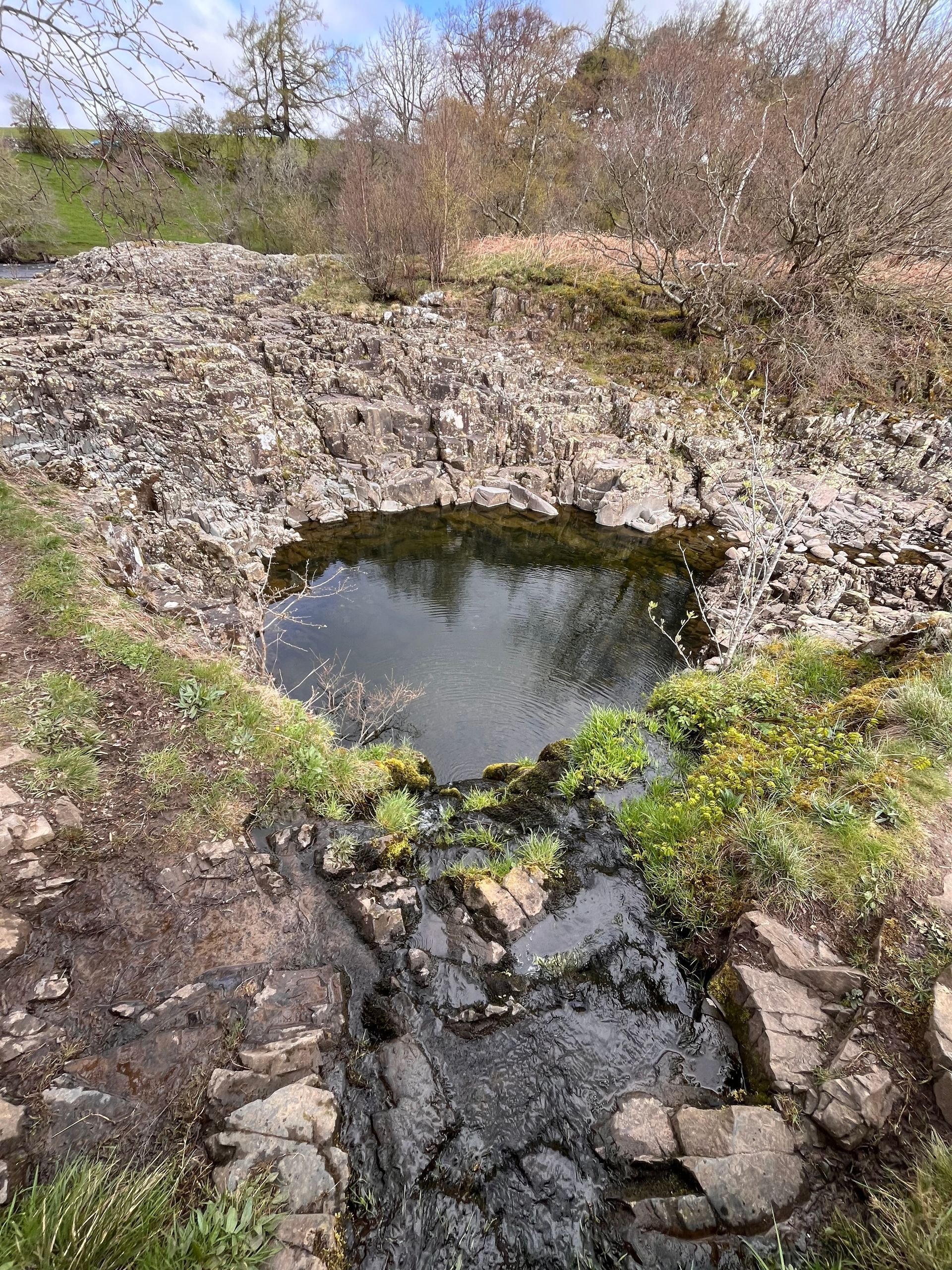 A small, dark pool of water set in a rocky landscape with sparse vegetation under a bright, cloudy sky.