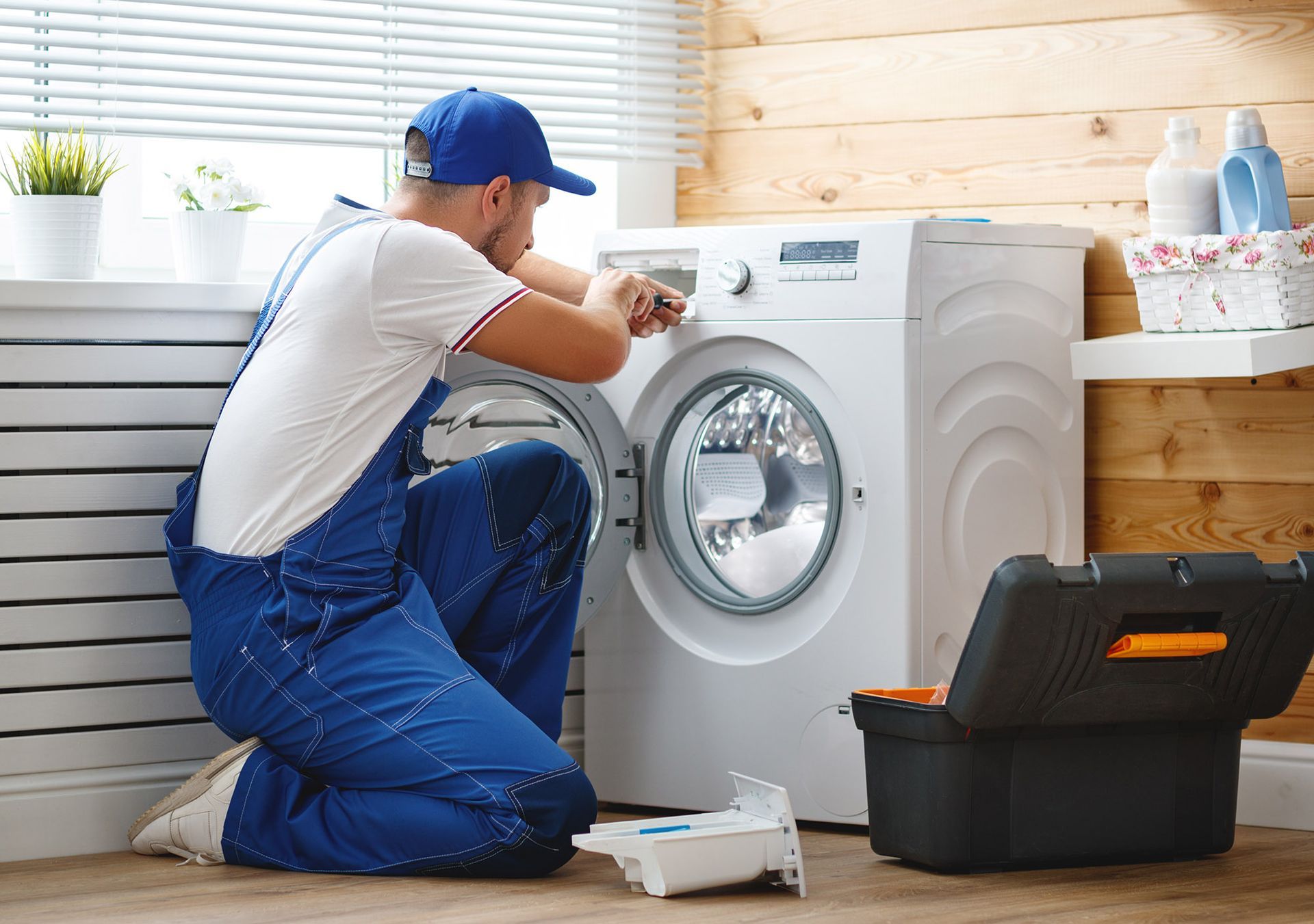 A person in blue overalls repairs a white washing machine in a laundry room with a toolbox.