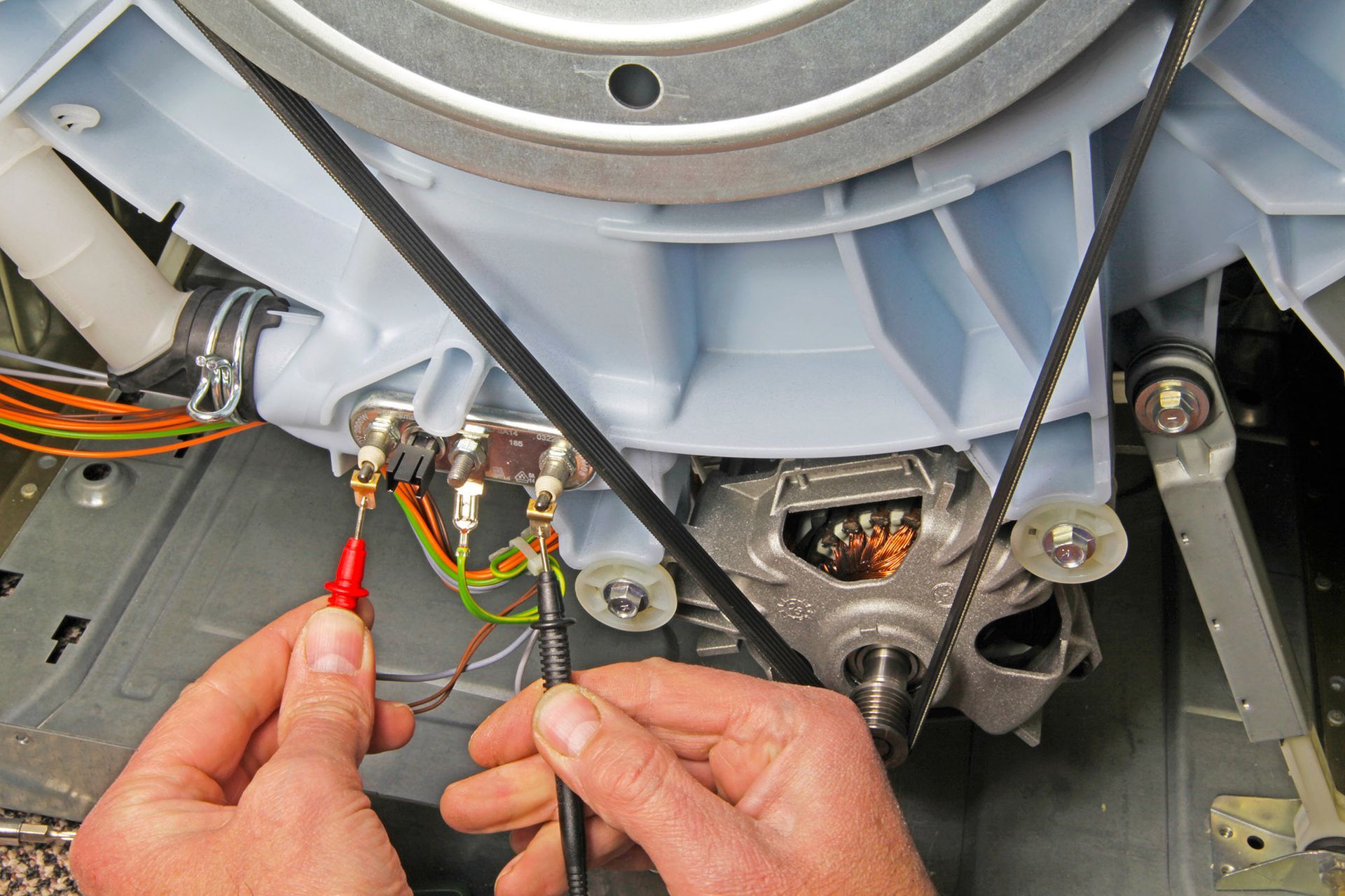 Hands testing electrical wires in a washing machine with a multimeter.