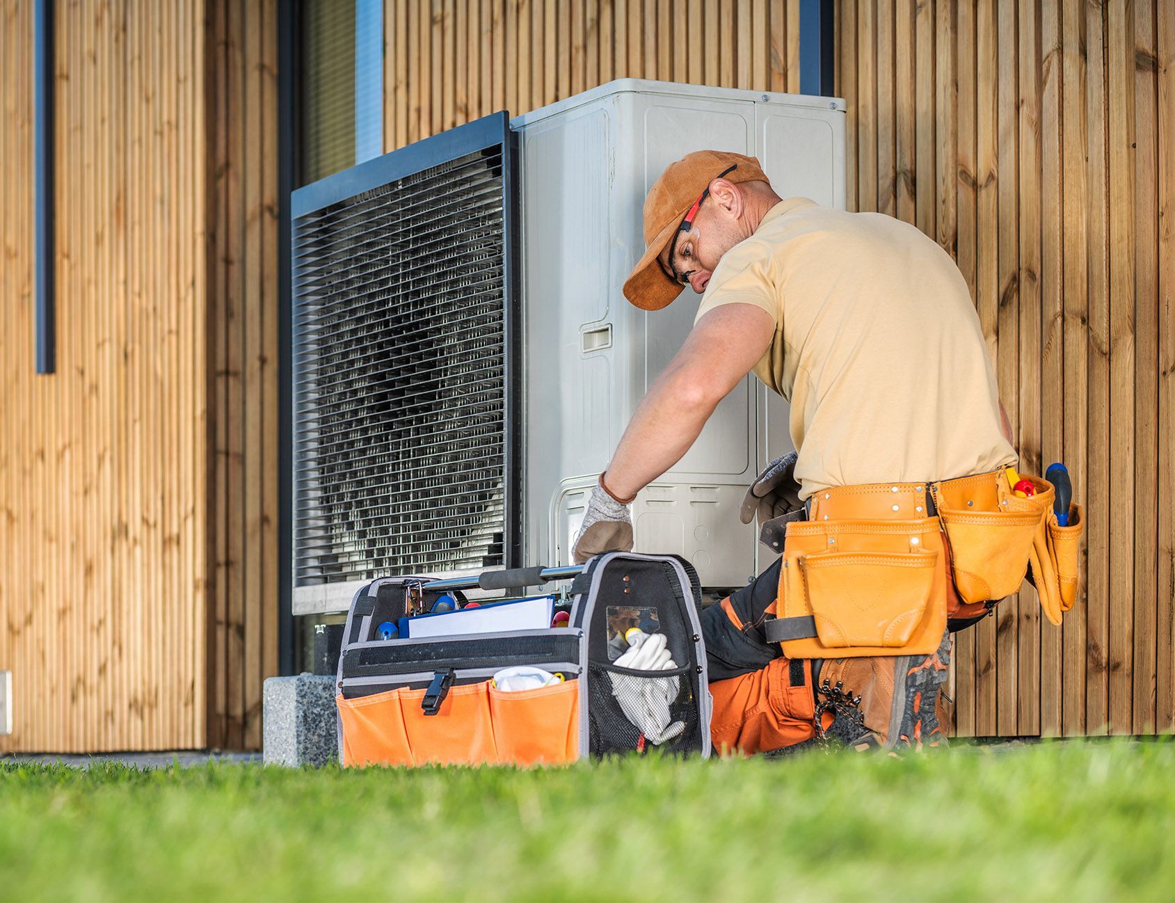 HVAC technician in a tan shirt and hat repairs an outdoor unit; tools and a toolbox nearby.