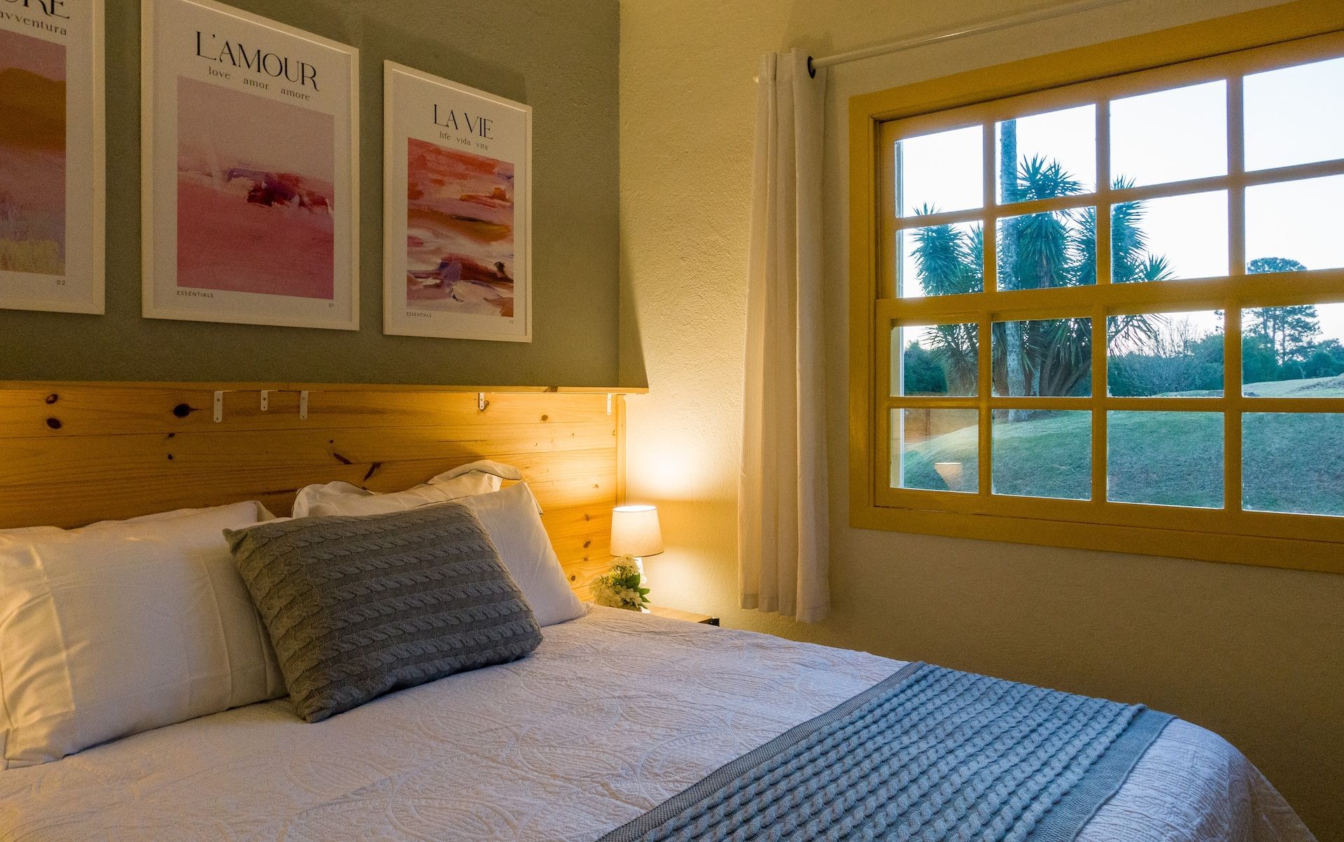 Quarto com cama, travesseiros, obras de arte emolduradas e uma janela com moldura amarela e vista para a vegetação.