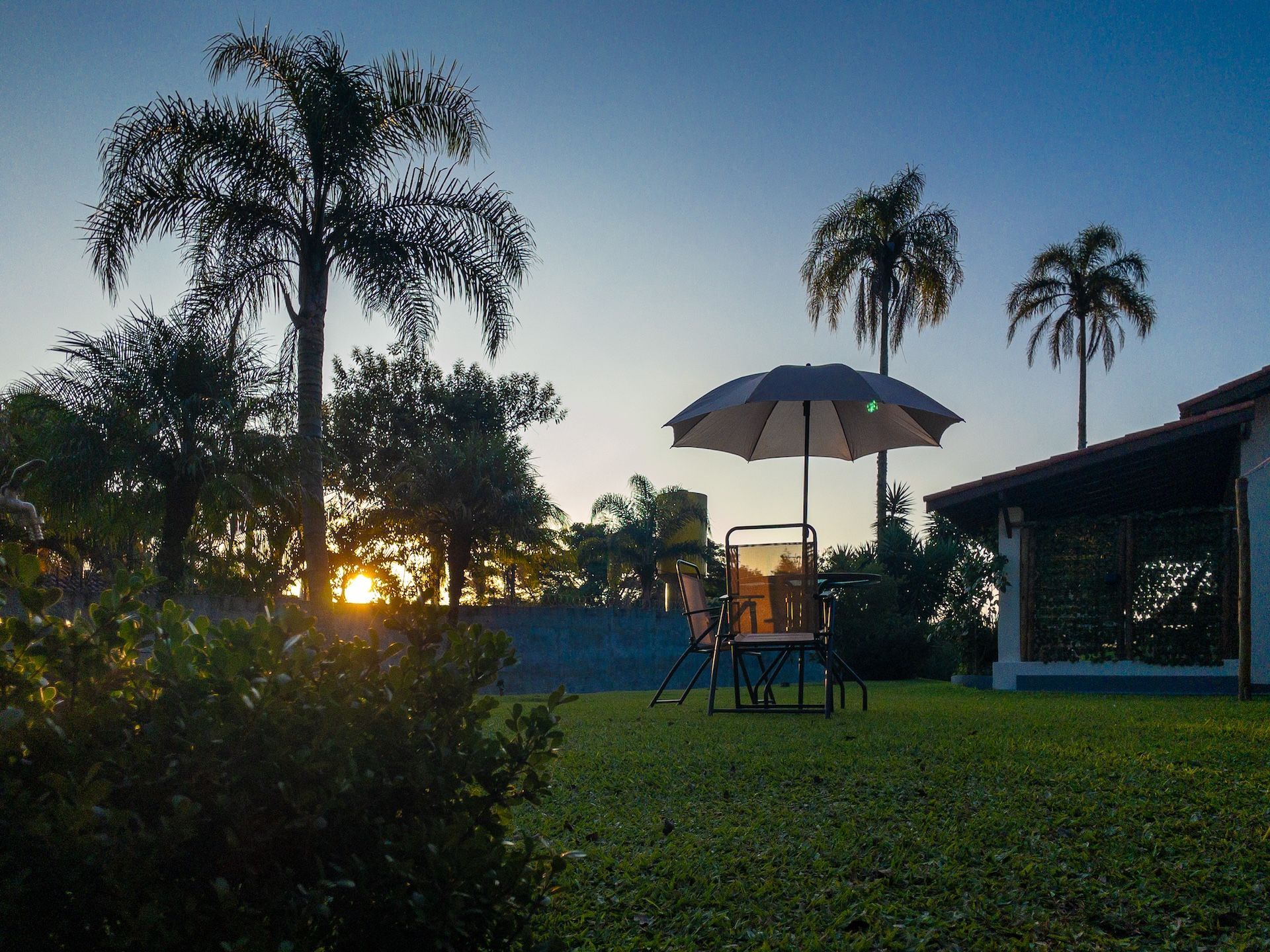 Pôr do sol sobre um gramado com árvores, um guarda-sol e uma cadeira perto de uma casa.