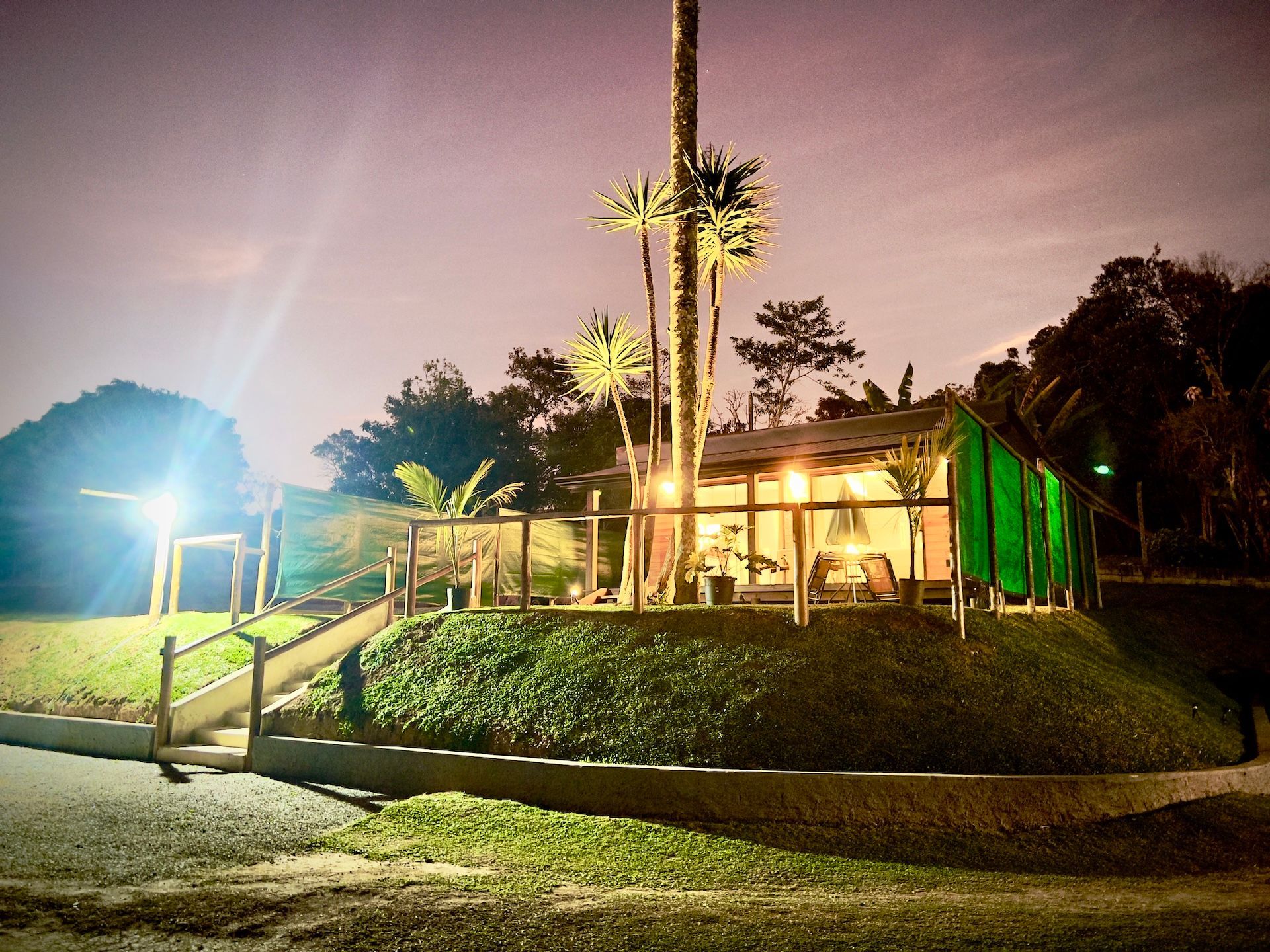 Vista noturna de uma pequena estrutura semelhante a uma cabana em uma colina gramada, iluminada por luzes externas. Palmeiras são visíveis.