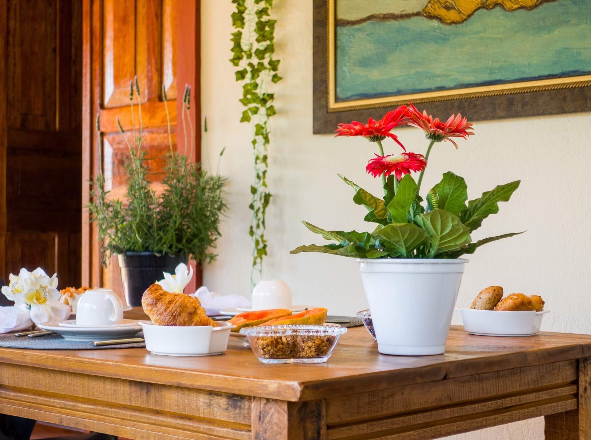 Mesa posta para o café da manhã com croissants, flores e um quadro.