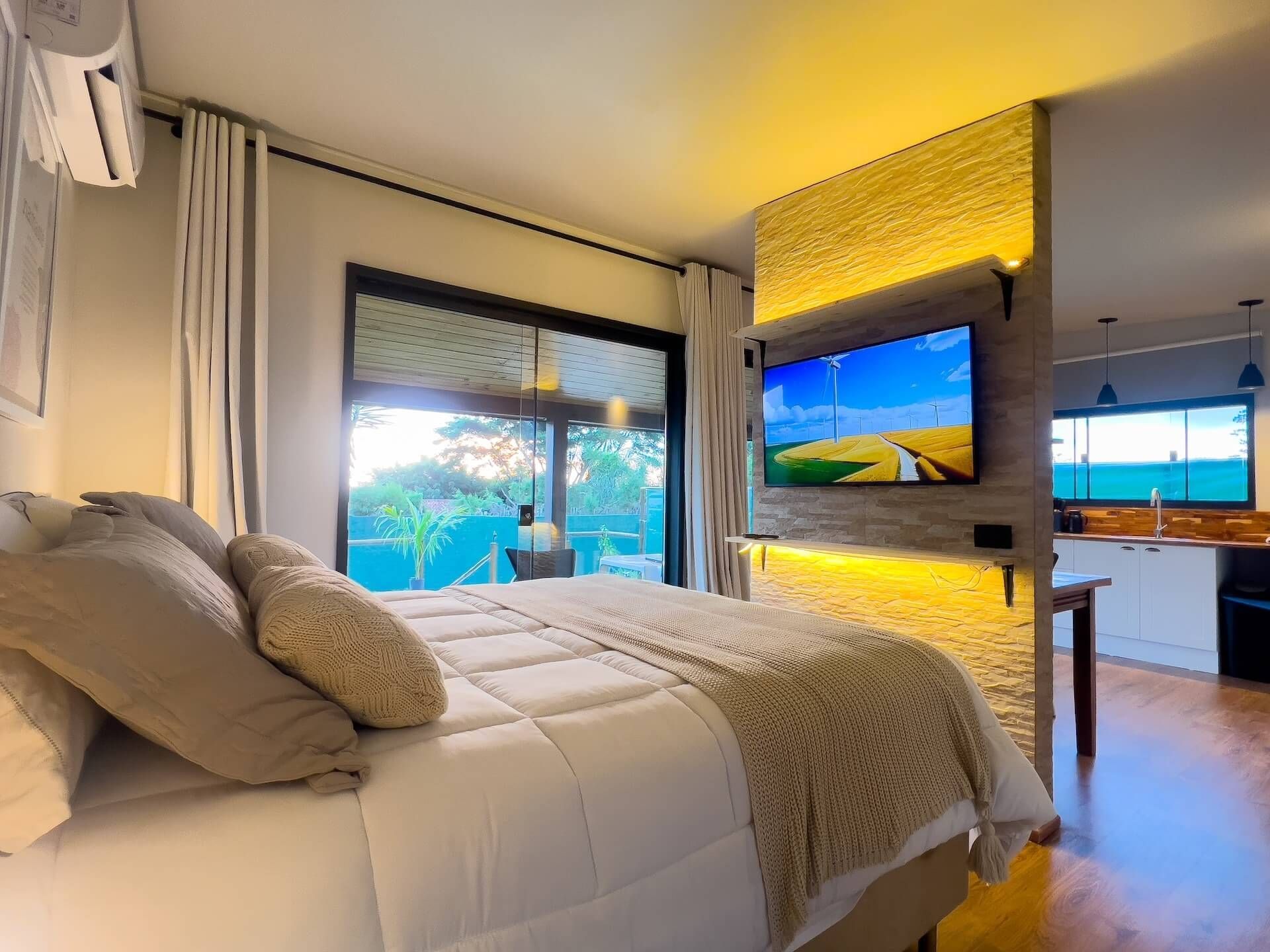 Quarto com cama, TV fixada na parede, portas abertas com vista para o exterior e área de cozinha adjacente.
