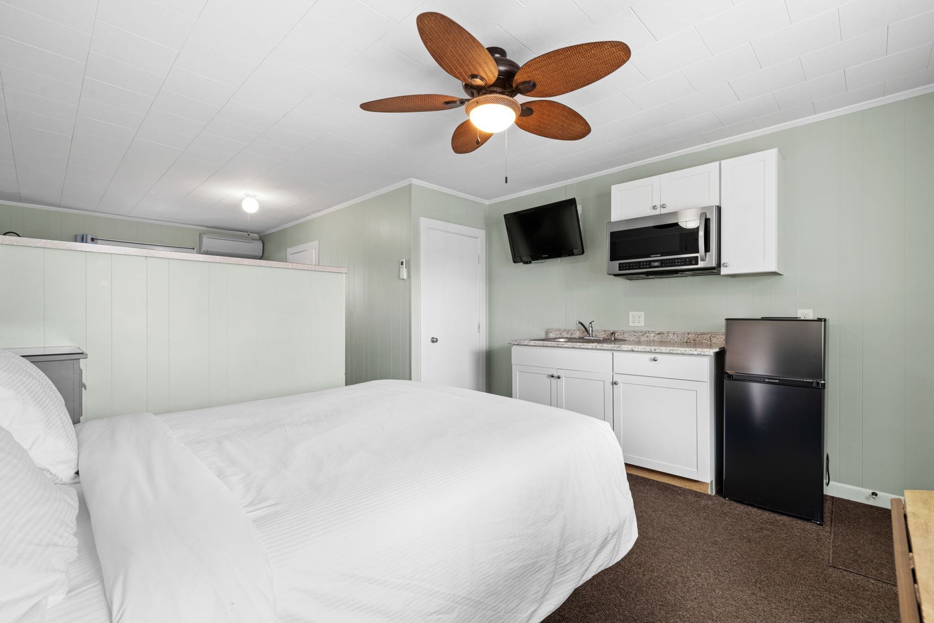 Small studio apartment with a bed, kitchenette, and TV; light green walls, brown carpet.