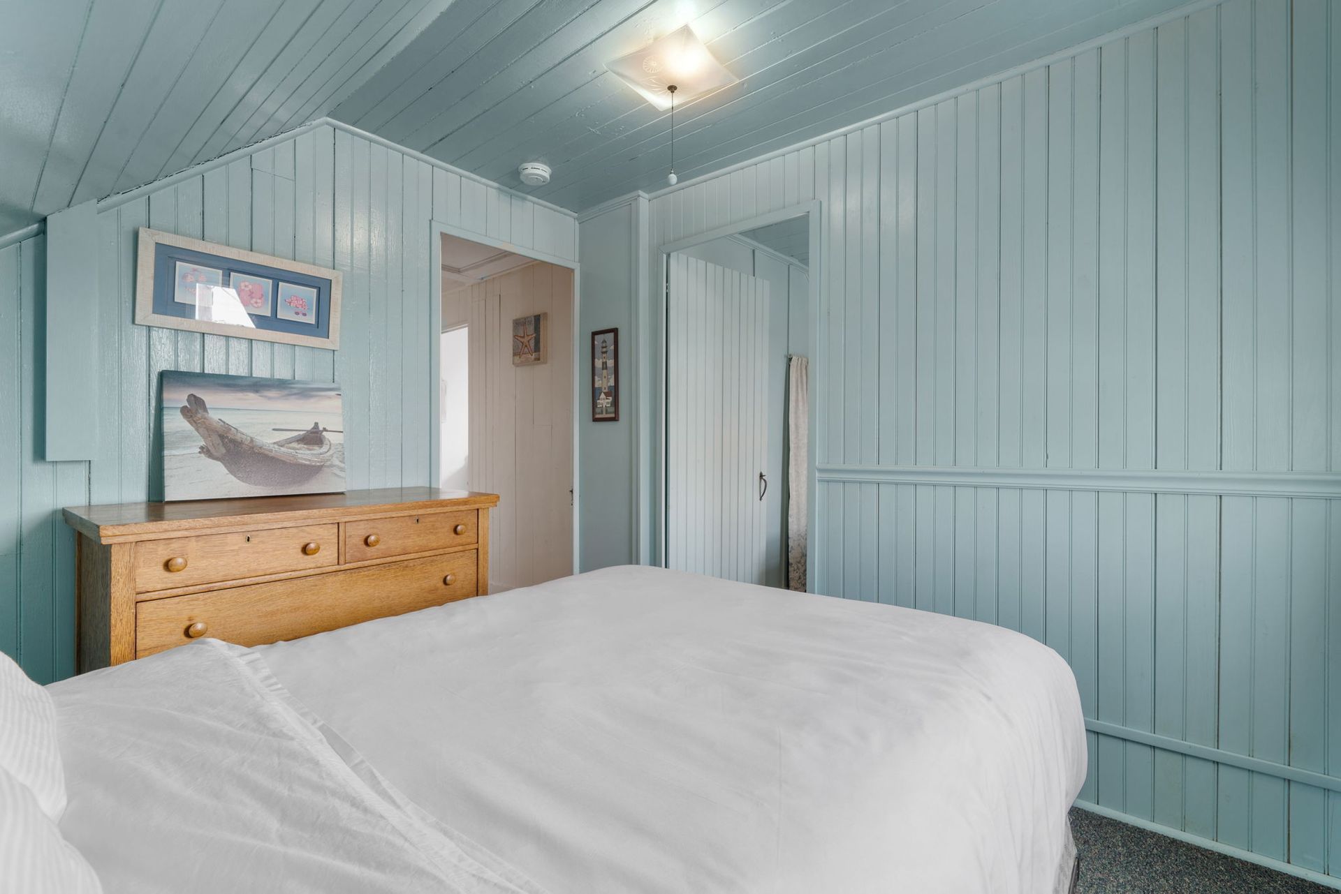 Bedroom with blue paneling, a light wood dresser, and a white bed.