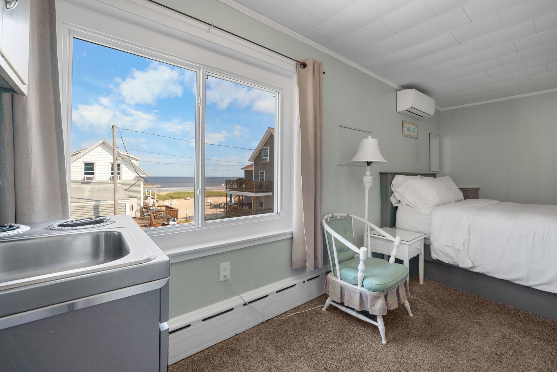 Small room with a bed, chair, sink, and large window overlooking the ocean and houses.