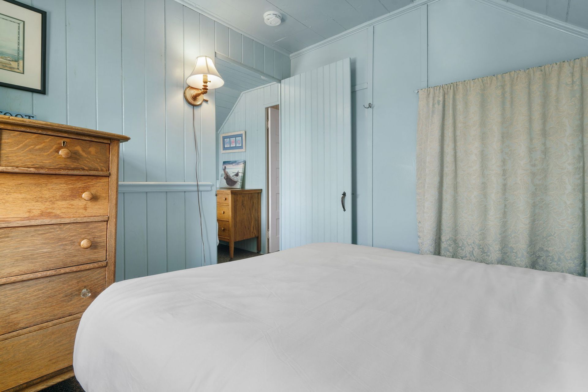 Bedroom with light blue paneled walls, a wooden dresser, and a bed with white linens.