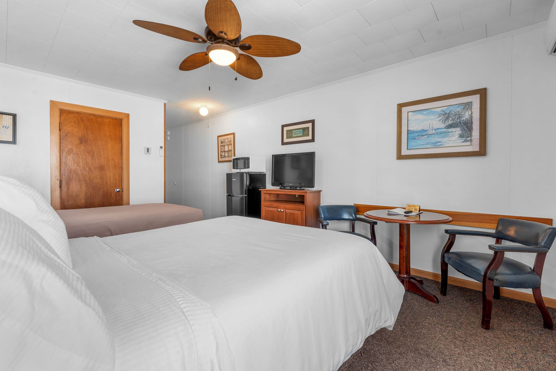 Bedroom with bed, small table & chair, TV, microwave, mini-fridge, and a door, white walls.