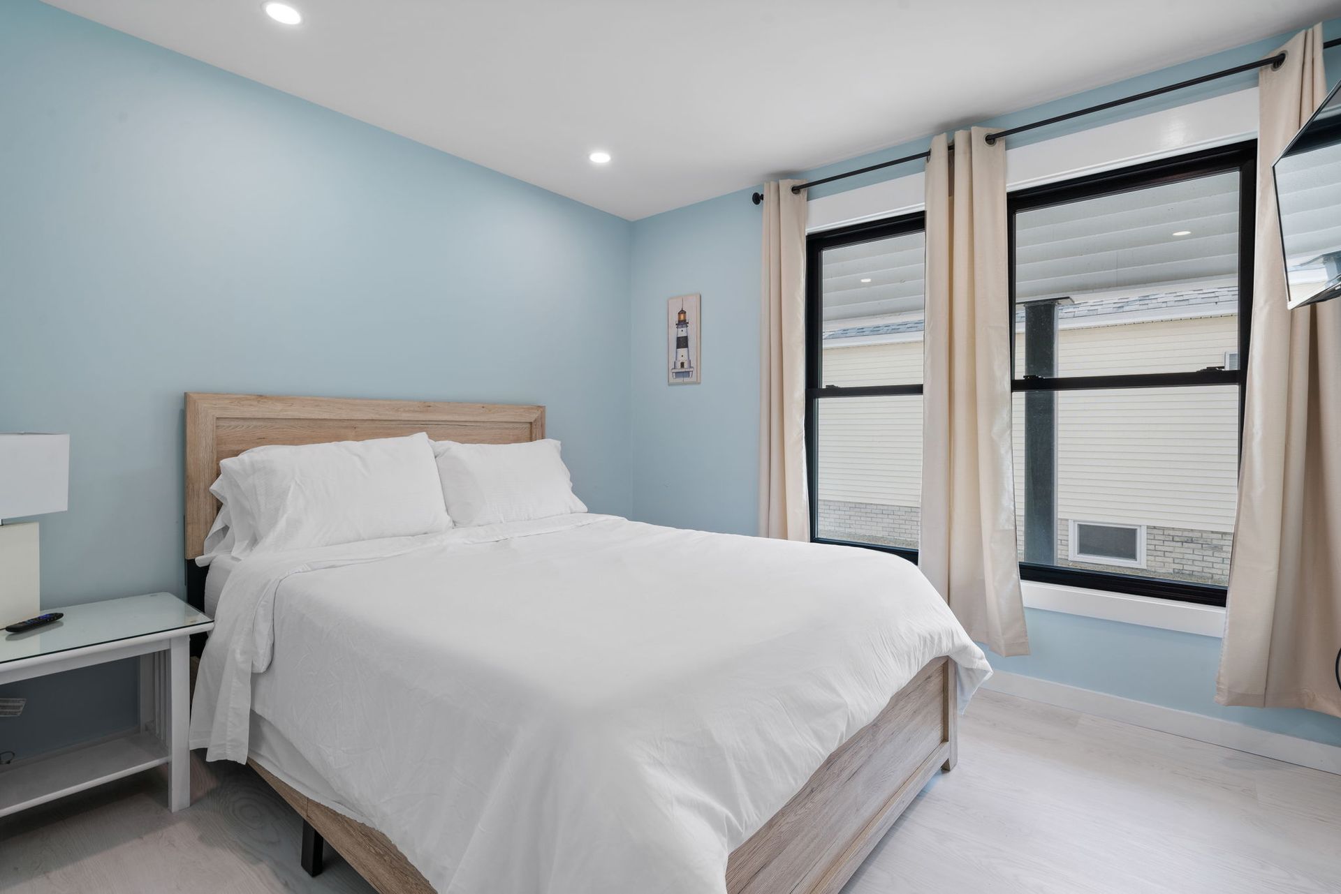 Bedroom with blue walls, white bedding, light wood bedframe, two windows with beige curtains.
