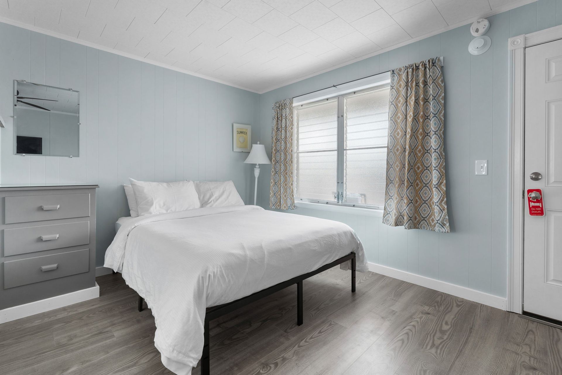 Bedroom with light blue walls, a bed, dresser, window with curtains, and a door.