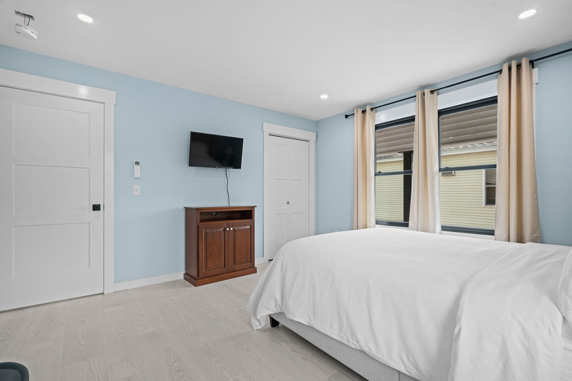 Bedroom with light blue walls, white bed, dark wood cabinet, and windows with beige curtains.