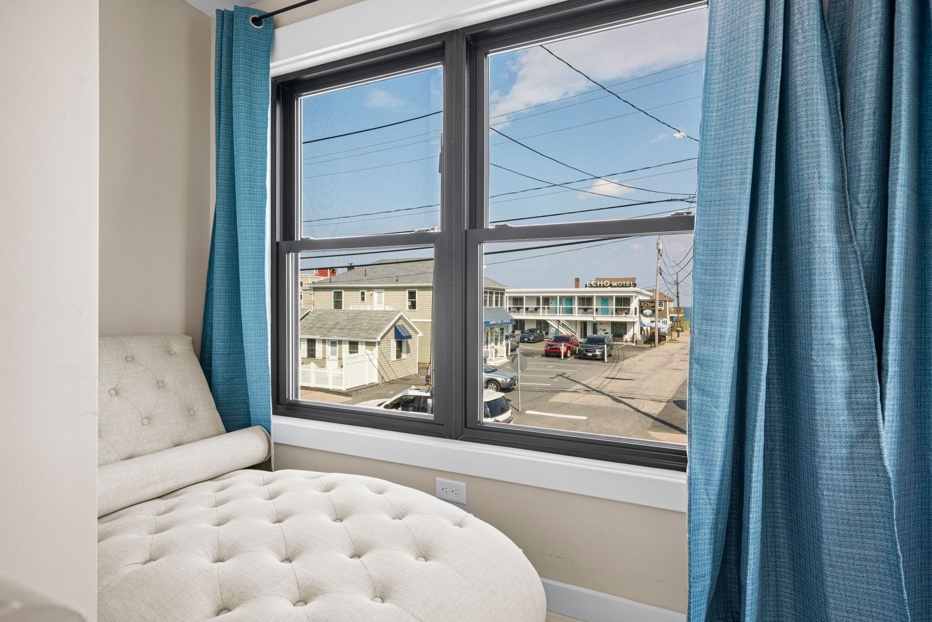 Window view with blue curtains, white tufted chaise lounge, and buildings in the background.