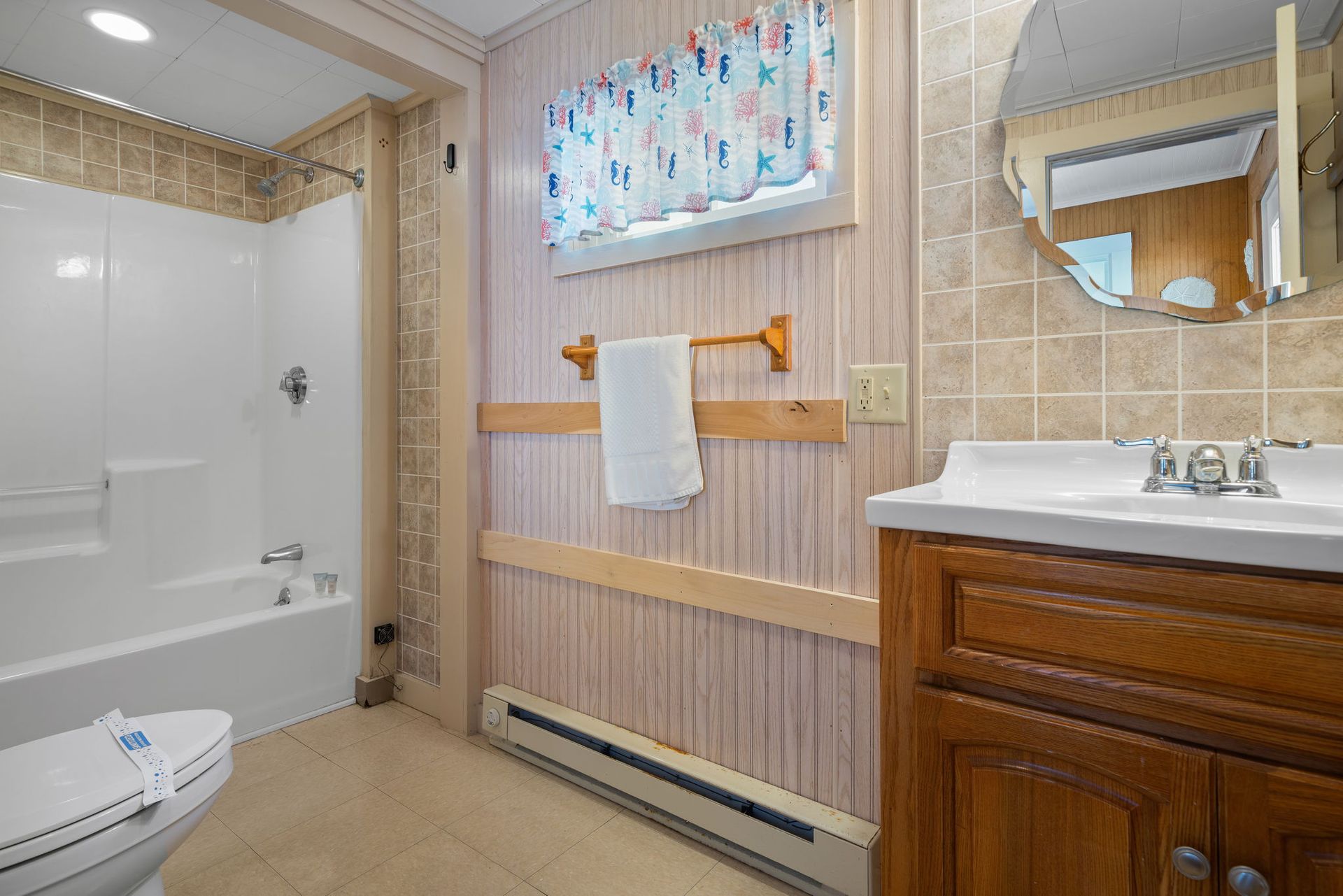 Bathroom with a toilet, bathtub, vanity, and a window with a seashell curtain.