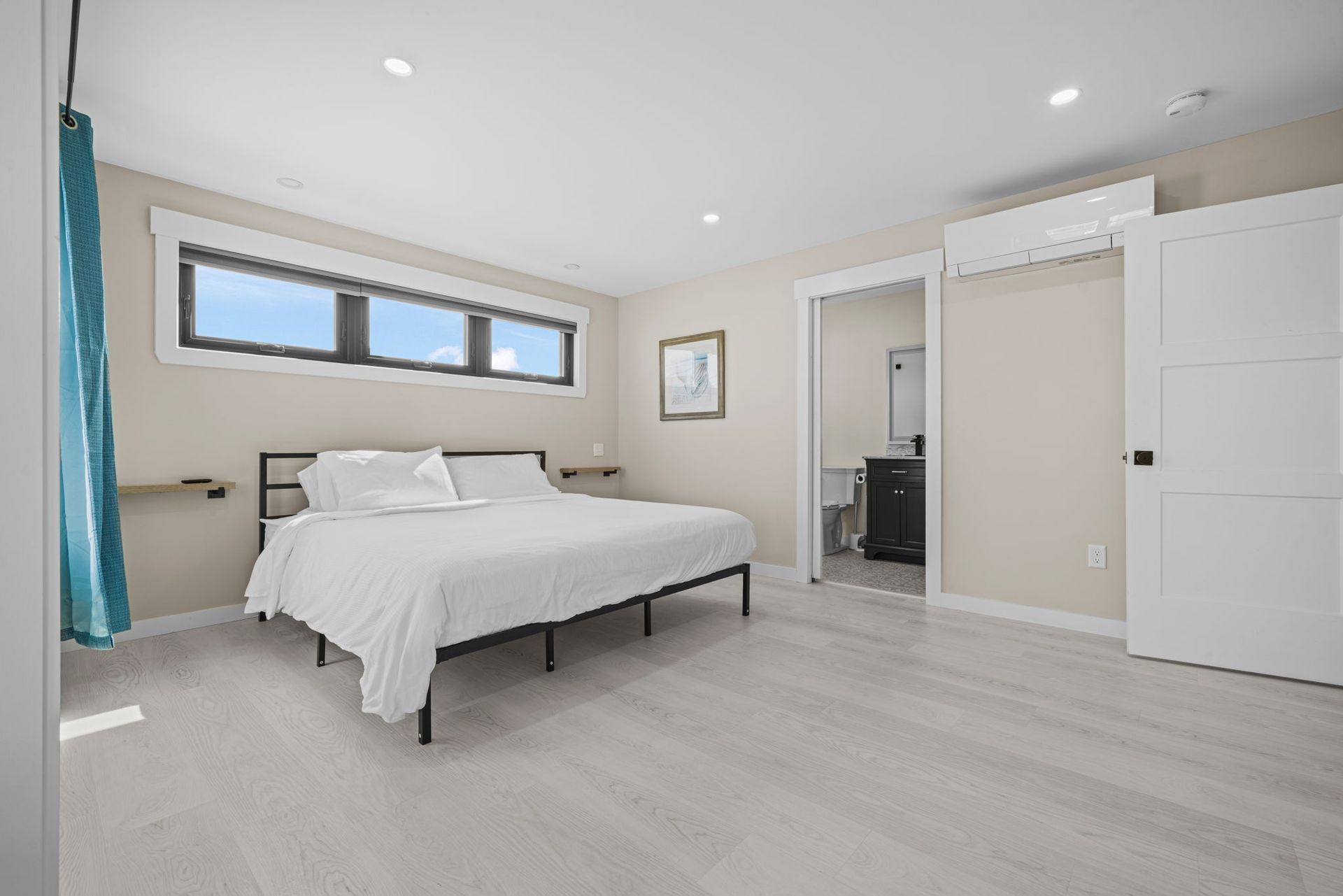 Bedroom with a white bed, light wood floors, a window, and an open door to a bathroom.