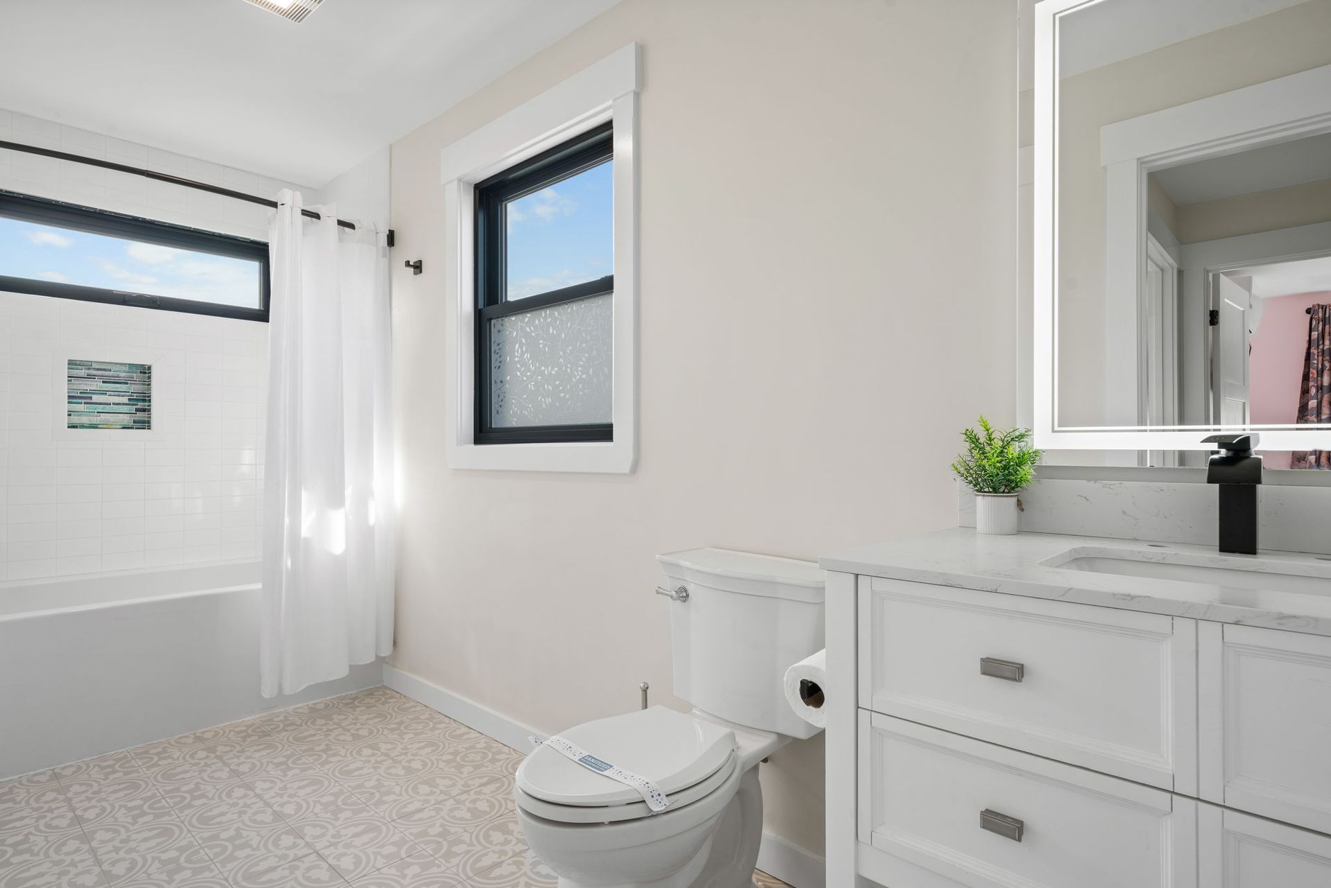 White bathroom with toilet, vanity, tub, and window with a black frame.