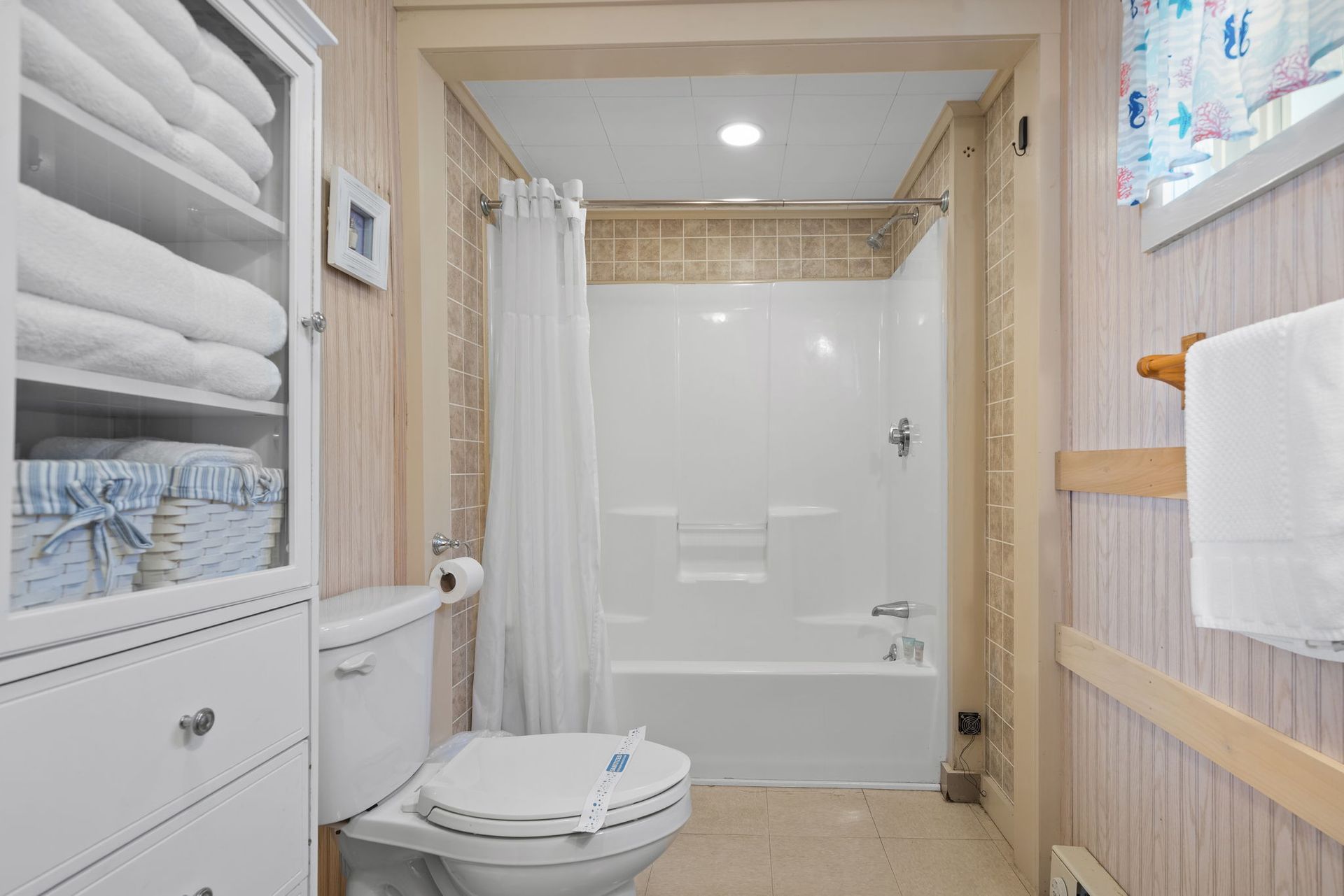 Bathroom with white toilet, shower, towels, and shelves. Light wood and cream tones.