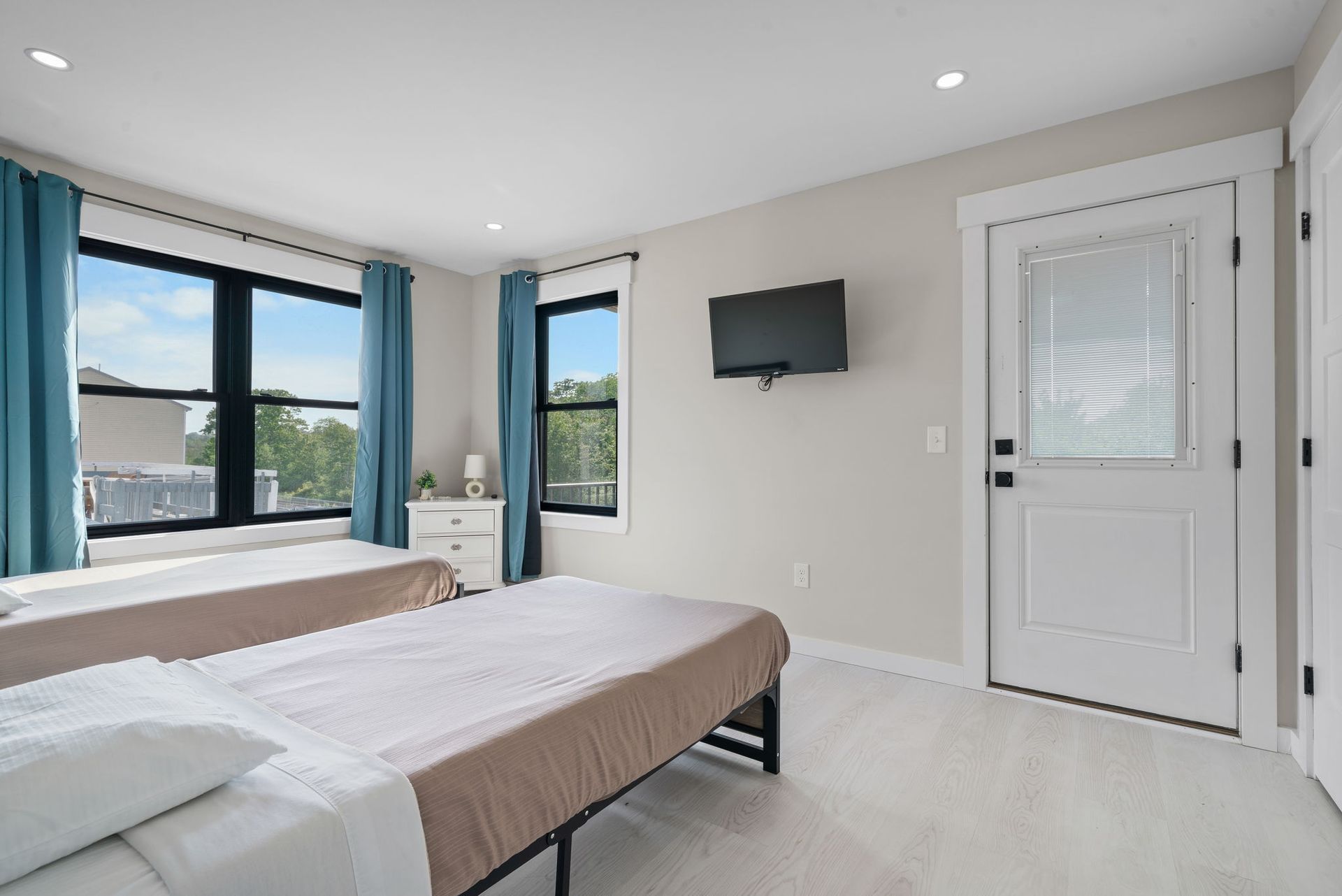 Bedroom with two twin beds, windows with blue curtains, white walls, and a mounted TV.