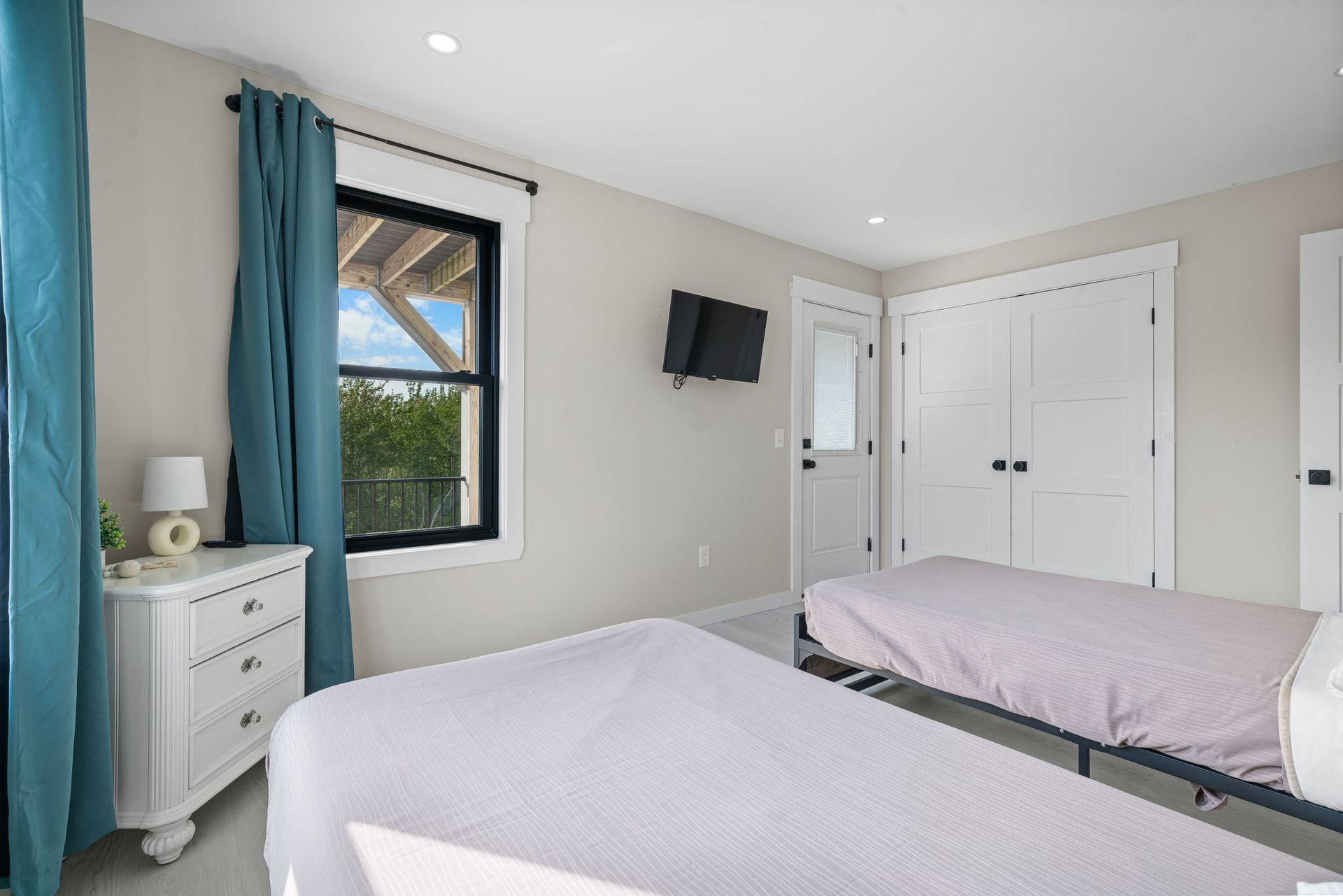 Bedroom with two beds, white dresser, teal curtains, window, and TV on the wall.