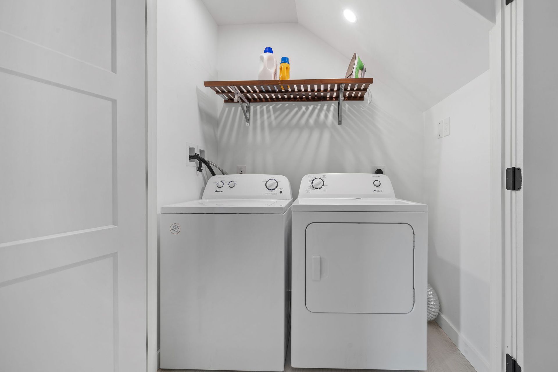 White washer and dryer in a small laundry room with a wooden shelf holding laundry supplies.