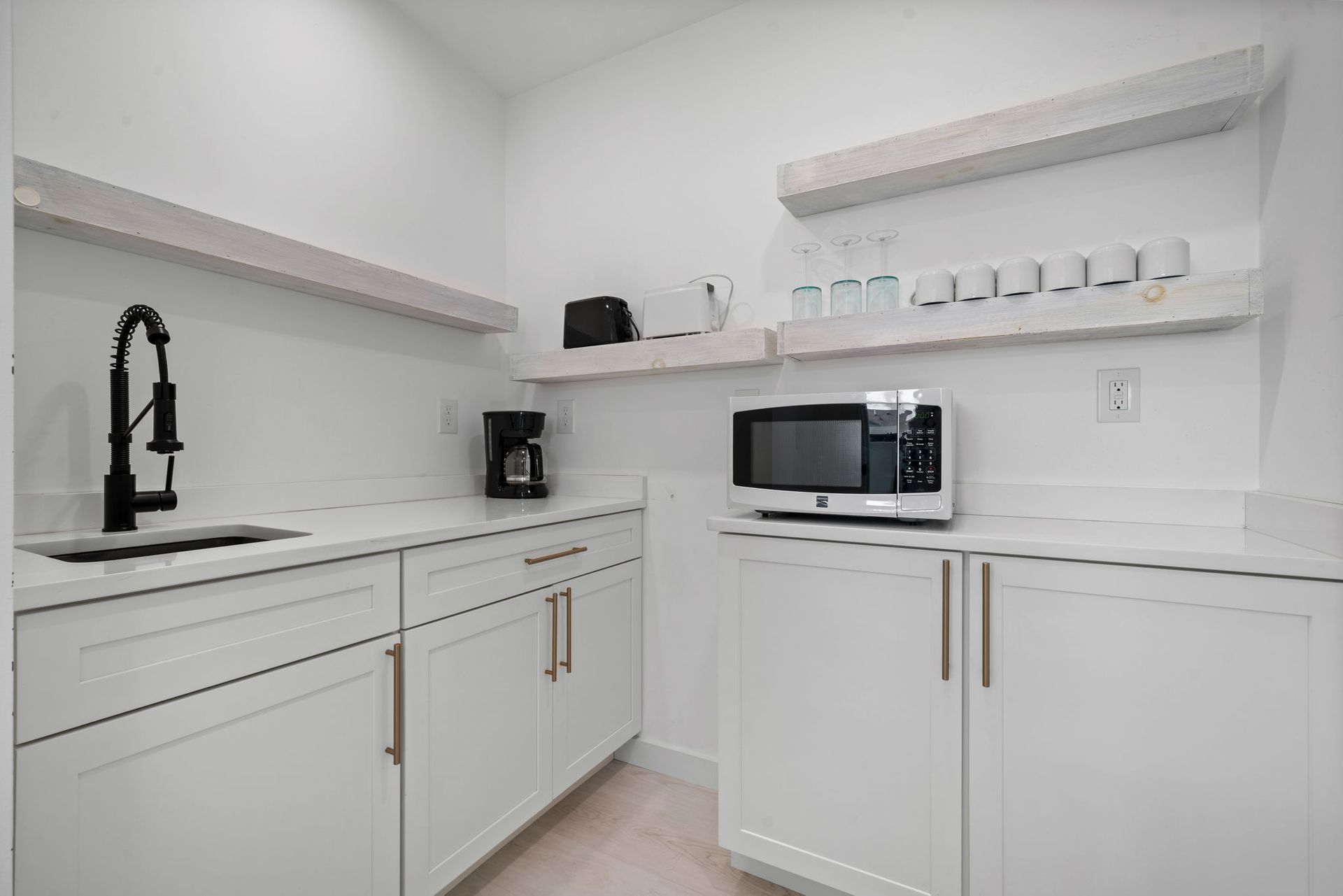 White kitchenette with sink, microwave, coffee maker, and floating shelves.