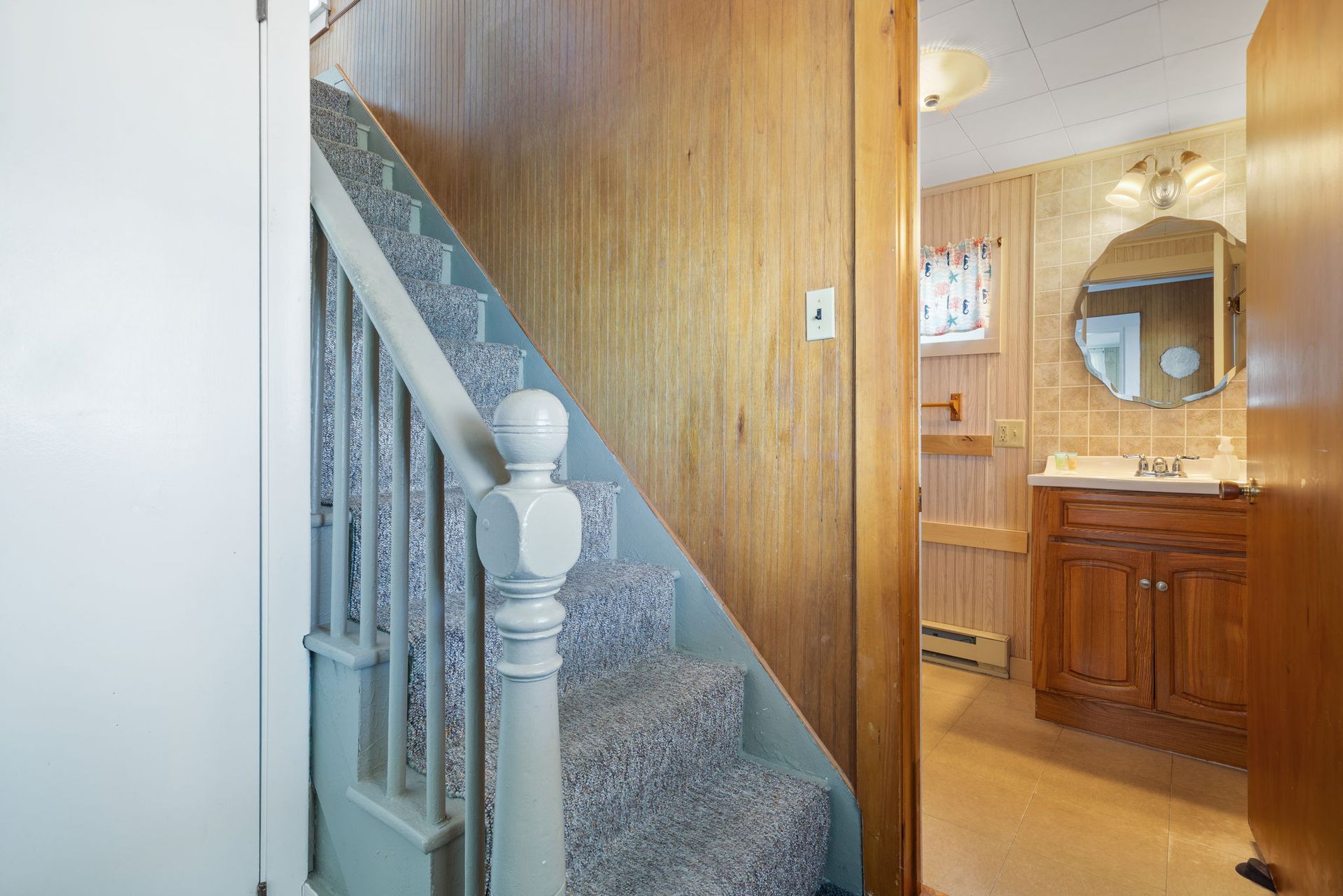 Staircase with gray carpet and blue banister leads to a bathroom with a wooden vanity and mirror.