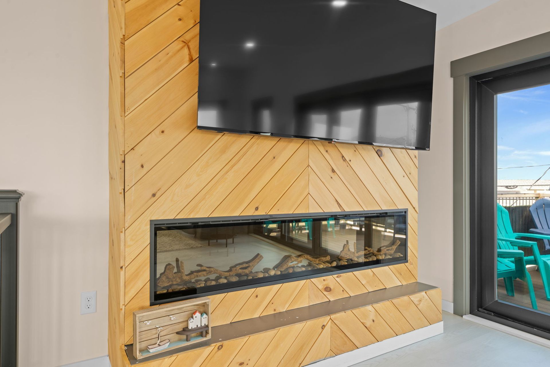 Fireplace and TV on a light wood accent wall. A sliding glass door reveals a deck with blue chairs.