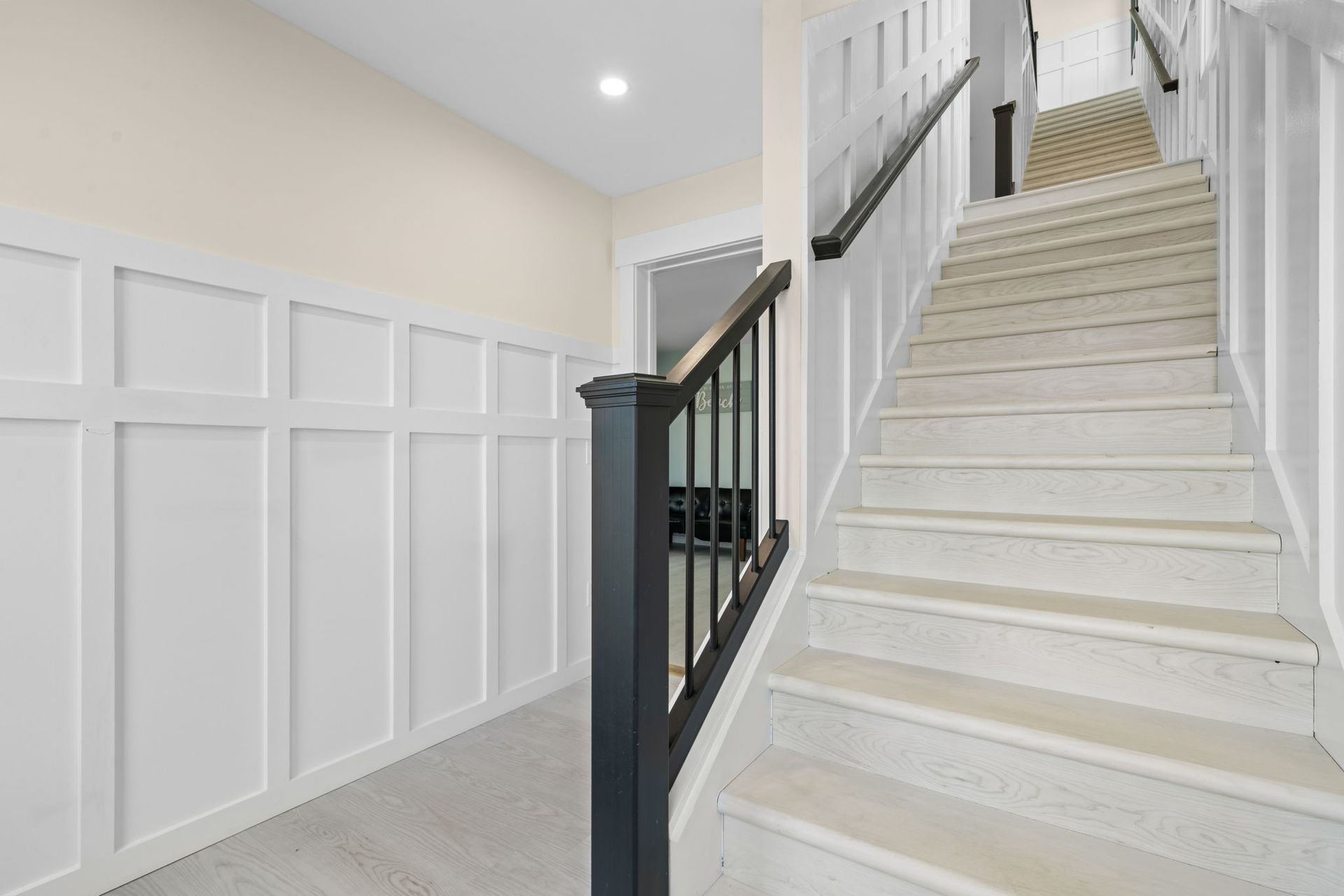 Hallway with white paneled wall, staircase with black railing, cream walls, and light wooden floors.