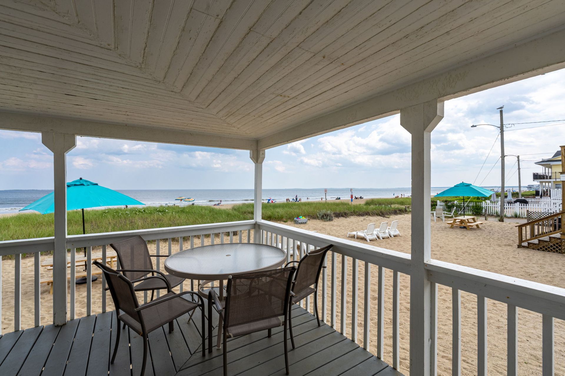 Porch overlooking a sandy beach with a table and chairs; teal umbrellas in the distance.