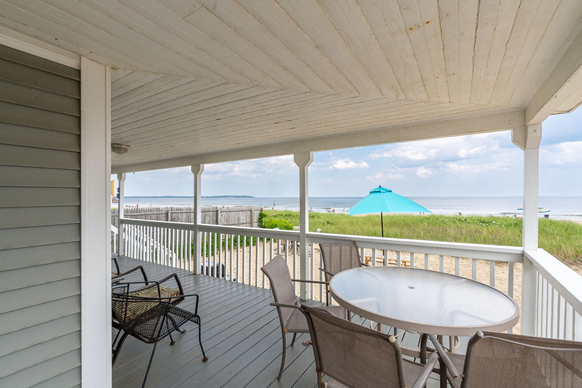 Covered porch overlooking a beach with chairs and a table, blue umbrella.