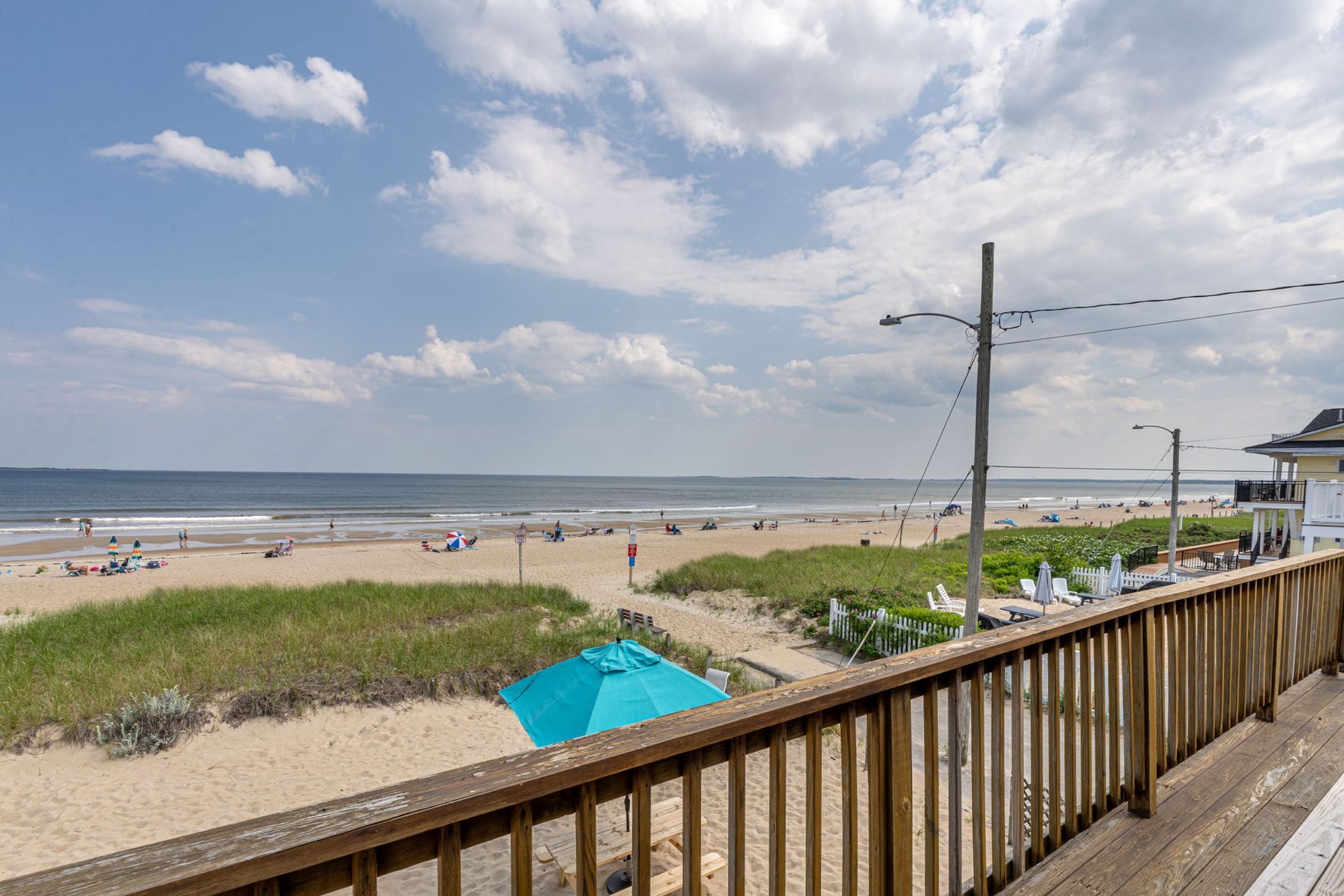 Beach view from a deck: blue umbrella, sandy beach, ocean, blue sky, people, and a wooden railing.