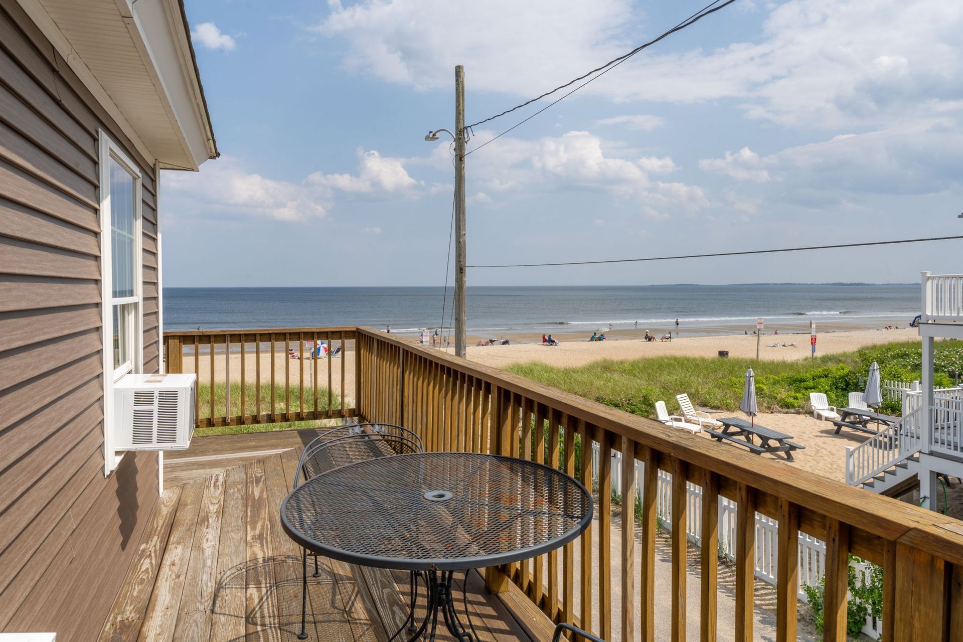Balcony overlooking beach with table, chairs, and ocean view. Brown building, blue sky.