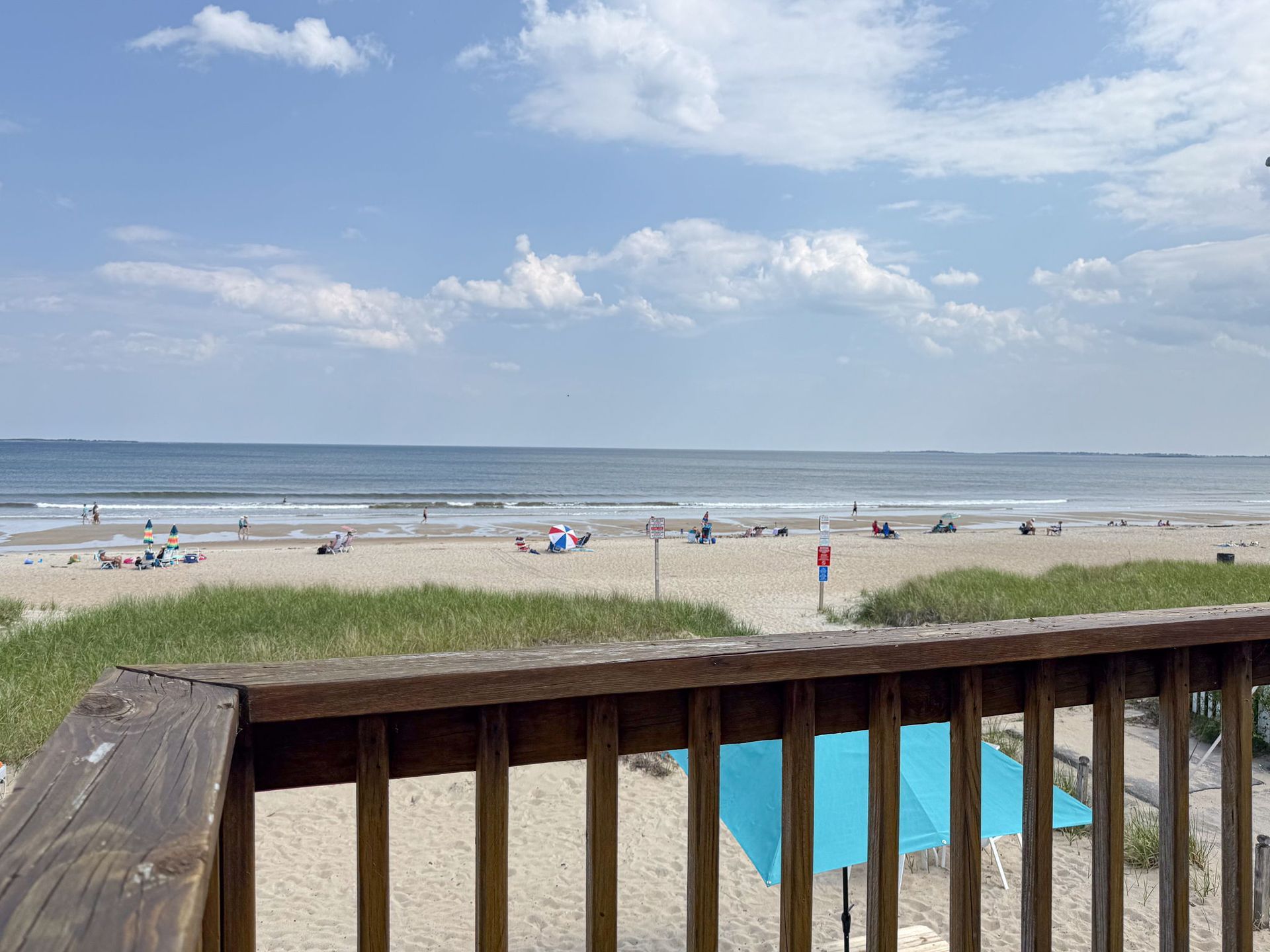 Beach scene with sand, ocean, and blue sky. View from a deck with a wooden railing. People are on the beach.