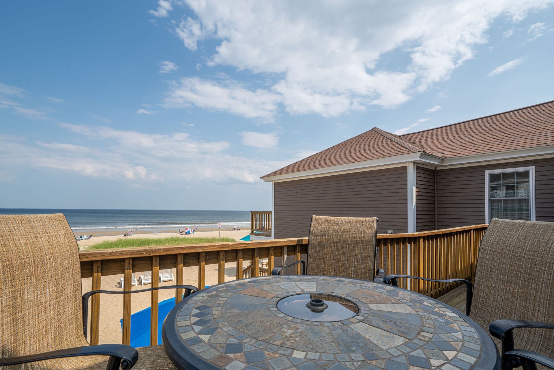 Deck with table and chairs overlooking beach and ocean under blue sky.