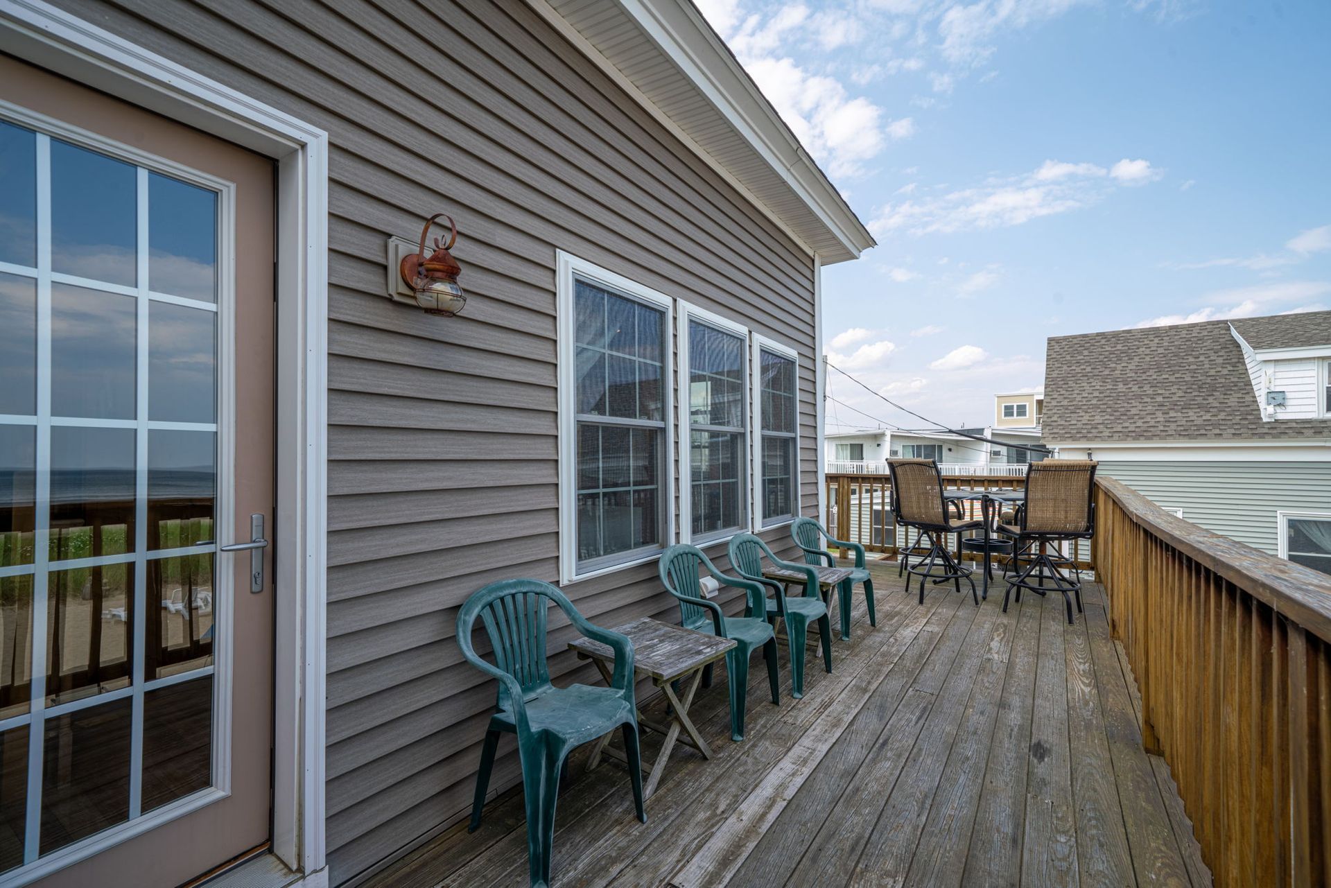Wooden deck with chairs, table, and beach view. Gray house siding, blue sky.